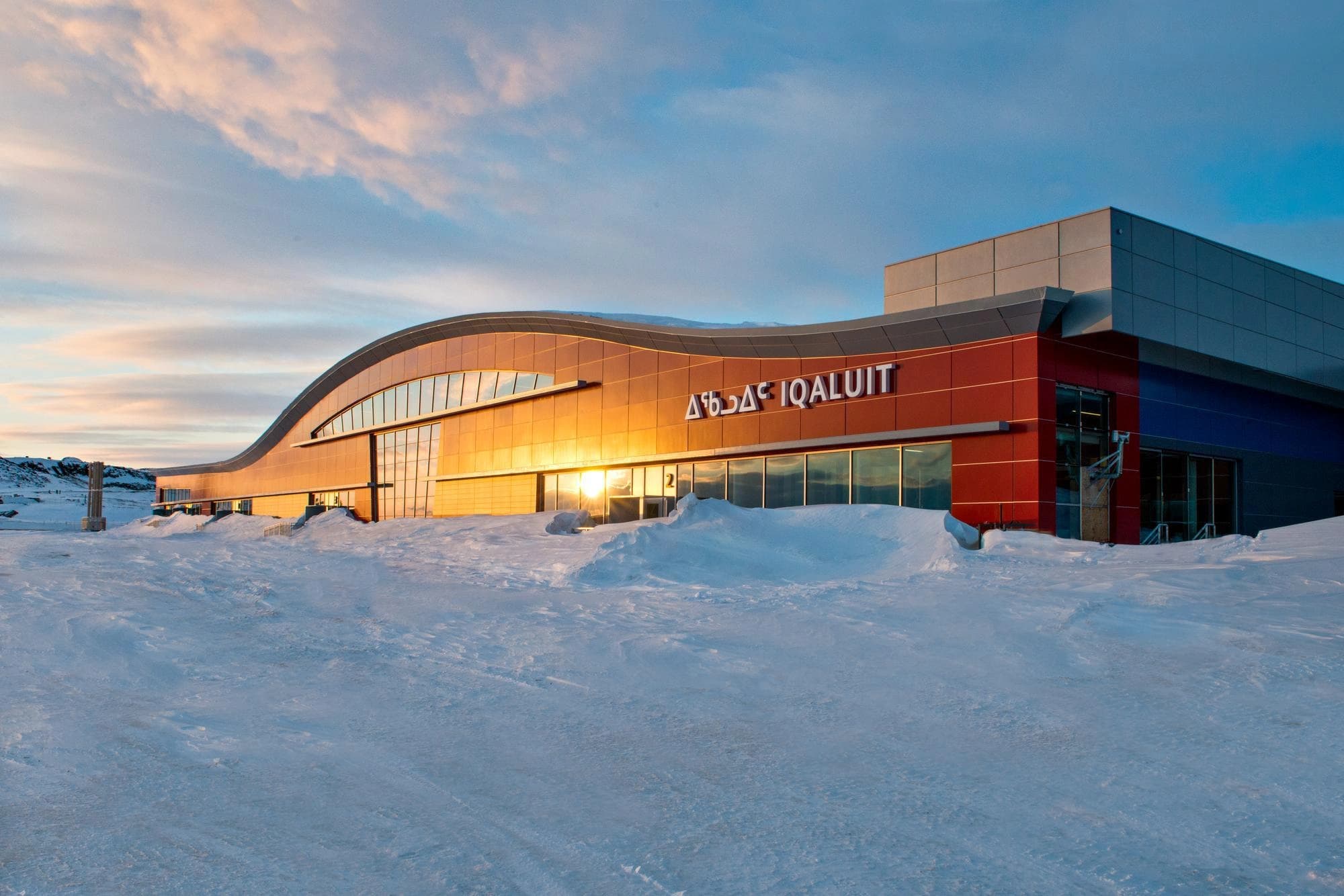 Le terminal moderne de l'aéroport d'Iqaluit, avec ses panneaux incurvés de verre et rouges, est baigné d'une lumière solaire dorée se reflétant sur sa façade. Entouré de neige blanche profonde, le panneau "ᐃᖃᓗᐃᑦ IQALUIT" est visible sous un ciel bleu et rose.