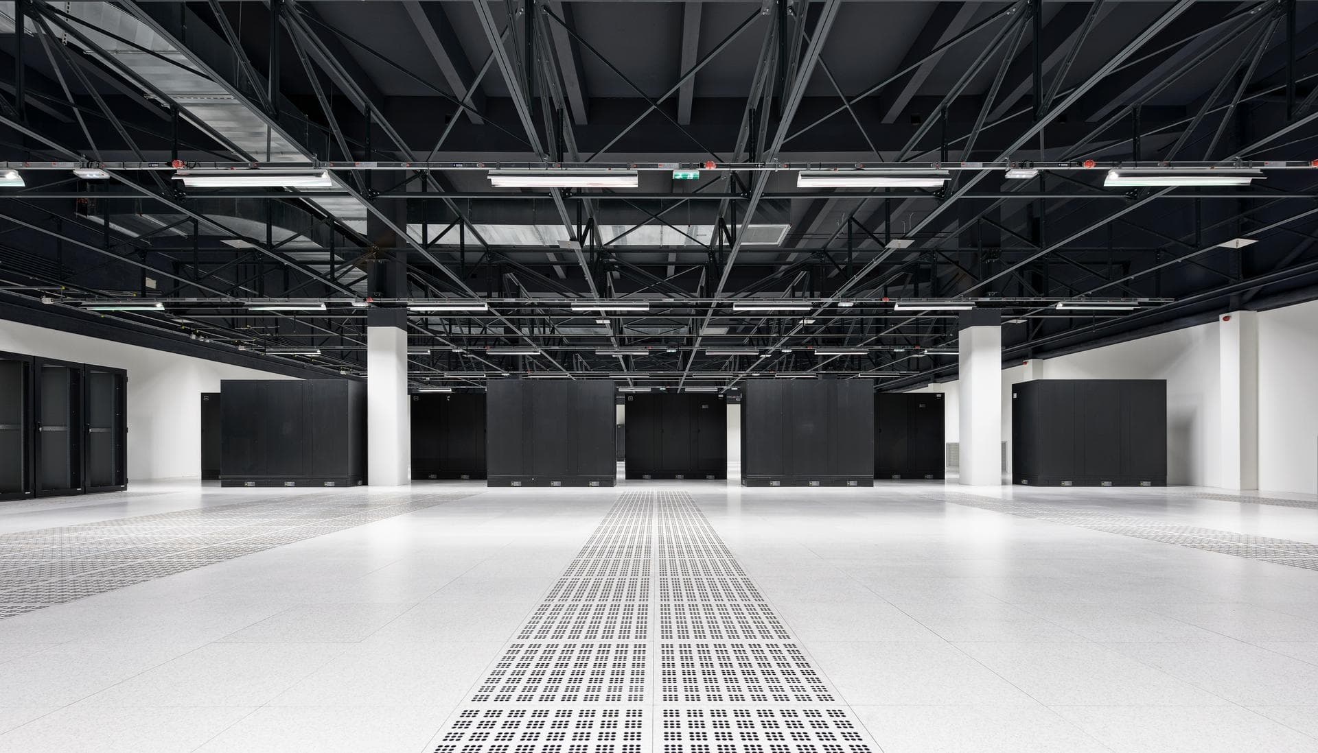Une vaste salle de centre de données avec un sol surélevé blanc et des rangées d'armoires de serveurs sombres sous un plafond industriel noir apparent équipé de nombreux luminaires.