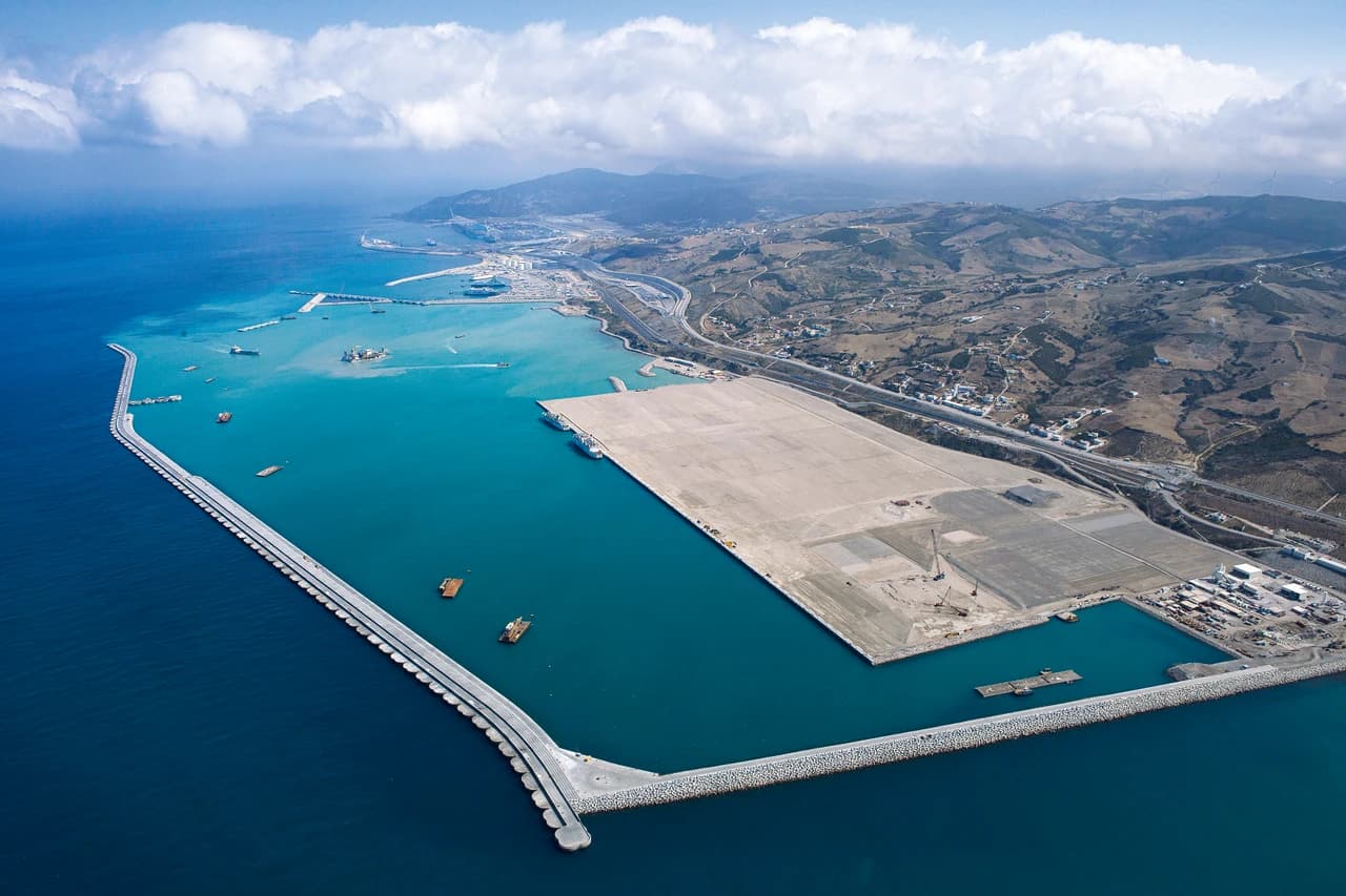 Vue aérienne d'un vaste port industriel moderne, avec un long brise-lames protégeant des bassins d'eau turquoise où flottent plusieurs navires et péniches, et d'immenses terre-pleins clairs pour les terminaux, le tout s'étendant au pied de collines arides sous un ciel parsemé de nuages.