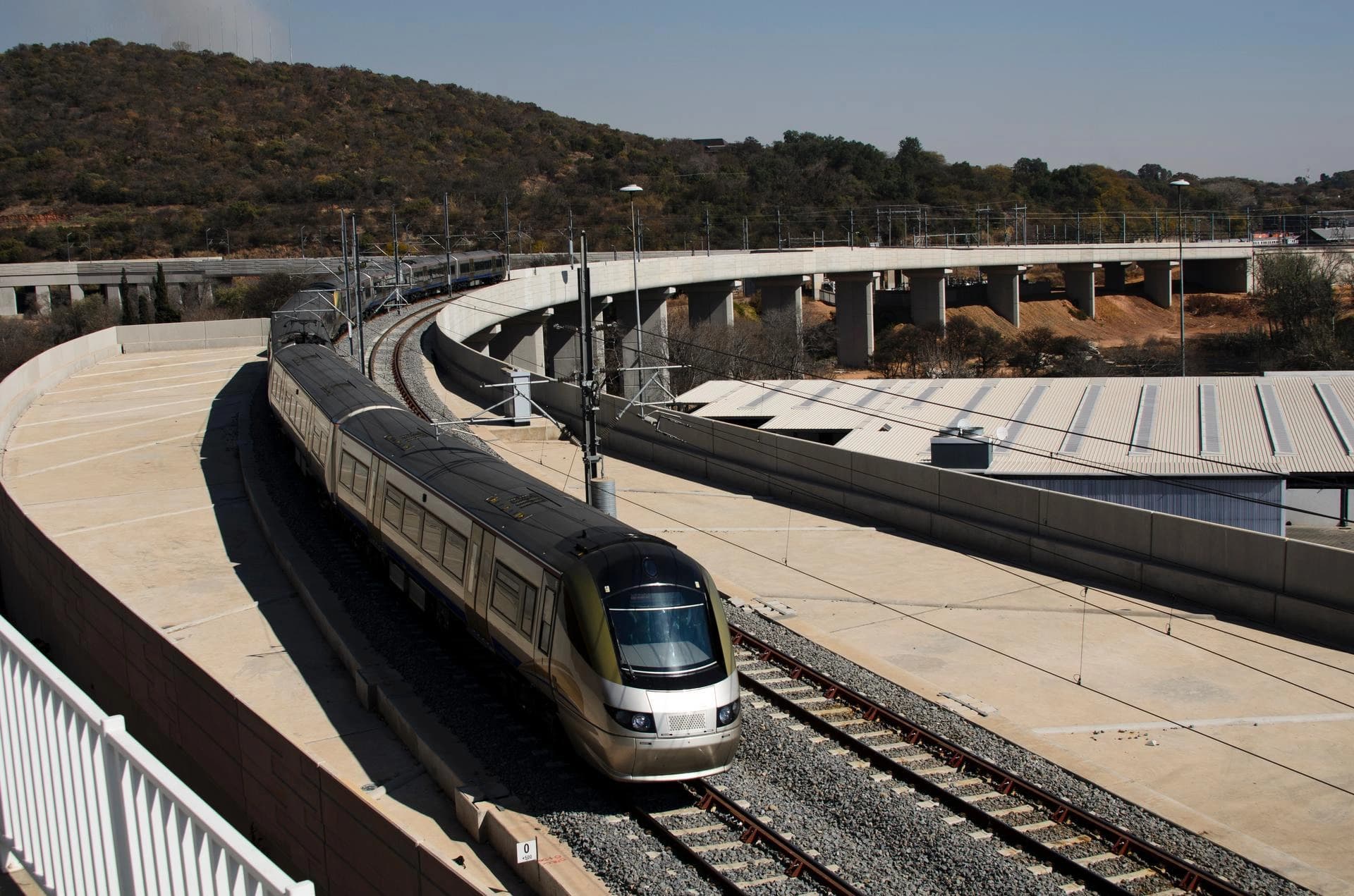 Un train moderne, élégant, aux couleurs or et noir, roule sur des rails courbés au premier plan, avec des ponts ferroviaires en béton et des collines arides sous un ciel clair en arrière-plan.