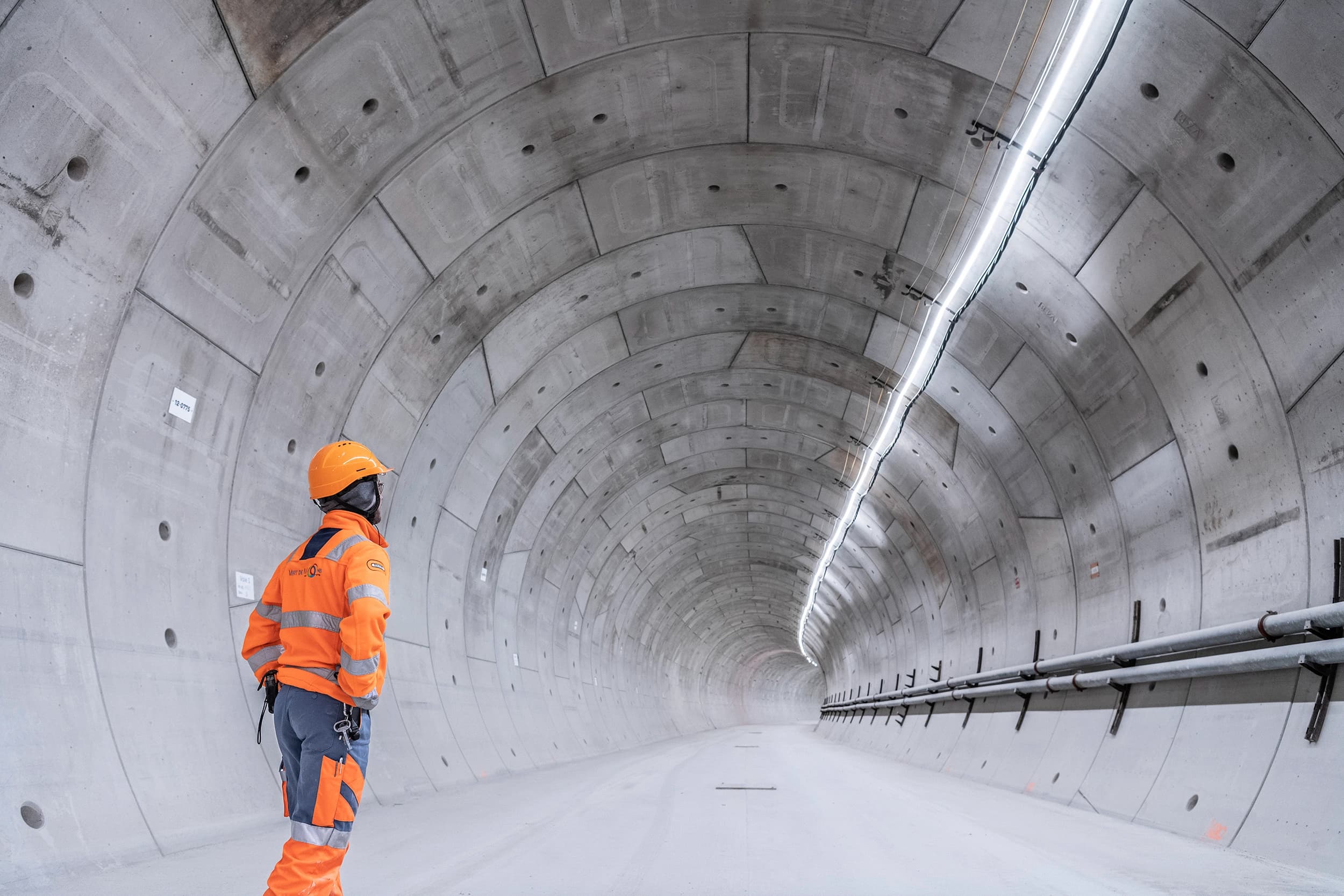 Un travailleur en tenue de sécurité orange vif et casque regarde dans un vaste tunnel en béton gris clair nouvellement construit, illuminé par une bande lumineuse LED.