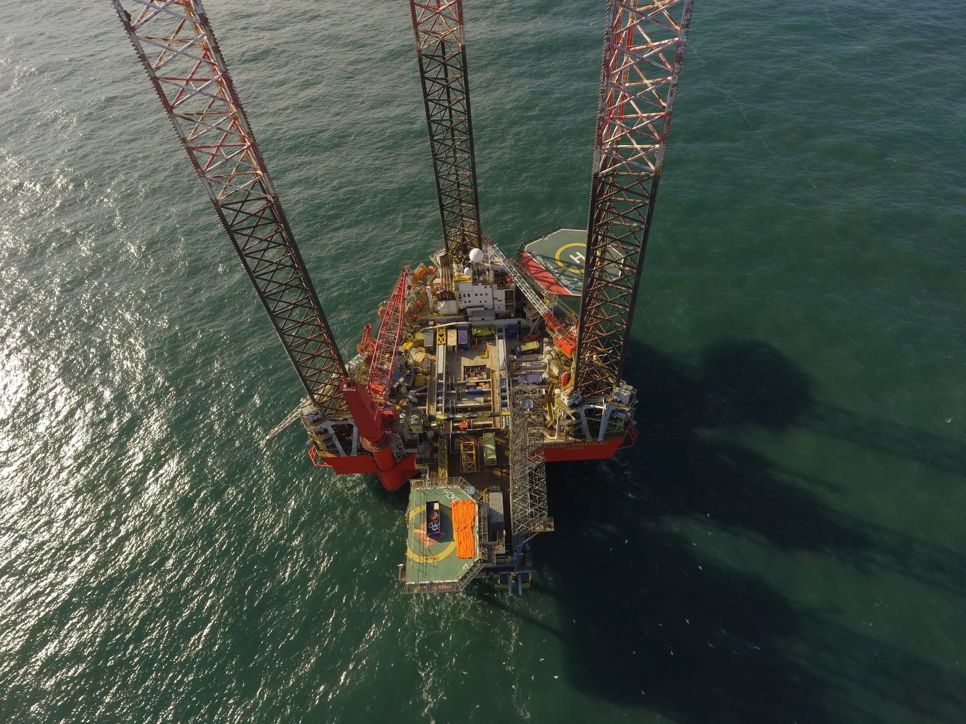 Vue aérienne d'une grande plate-forme pétrolière offshore rouge et métallique avec trois pieds massifs, ancrée dans une mer vert foncé brillante. Le pont de la plate-forme est détaillé avec divers équipements et un héliport marqué.