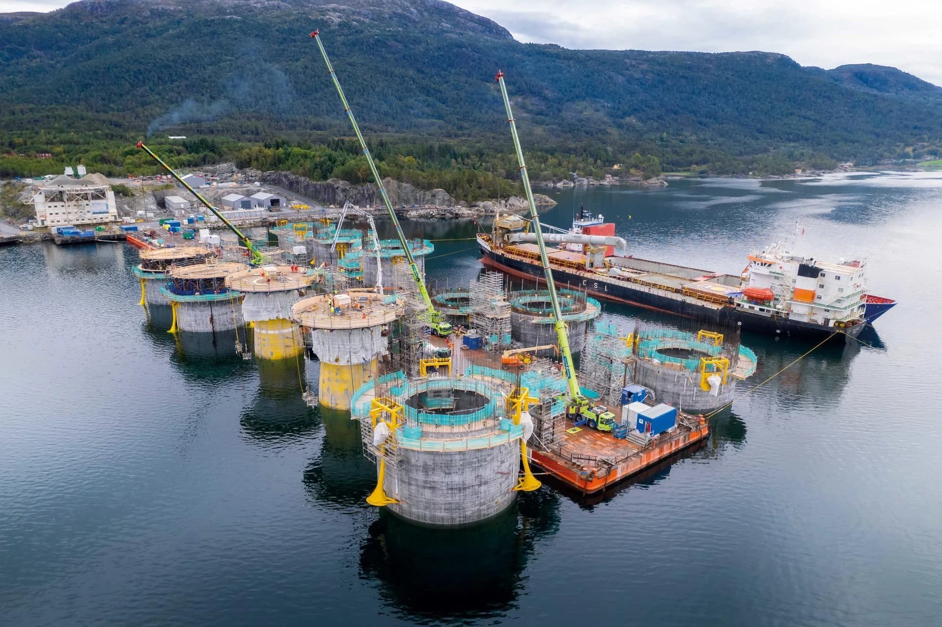 Vue aérienne d'un vaste chantier de construction dans un fjord, montrant de nombreuses structures cylindriques en béton grises et jaunes en cours d'assemblage par des grues et des échafaudages. Un grand cargo est amarré à proximité, avec des montagnes boisées en arrière-plan sous un ciel couvert.