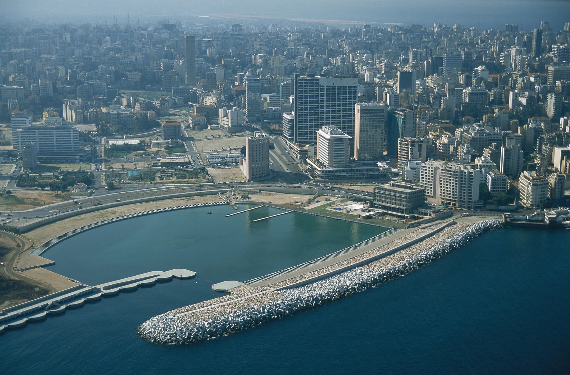 Vue aérienne d'une ville côtière dense sous un ciel brumeux, montrant de nombreux immeubles gris modernes. Au premier plan, une grande marina aux eaux turquoise est protégée de la mer d'un bleu profond par une longue digue rocheuse.