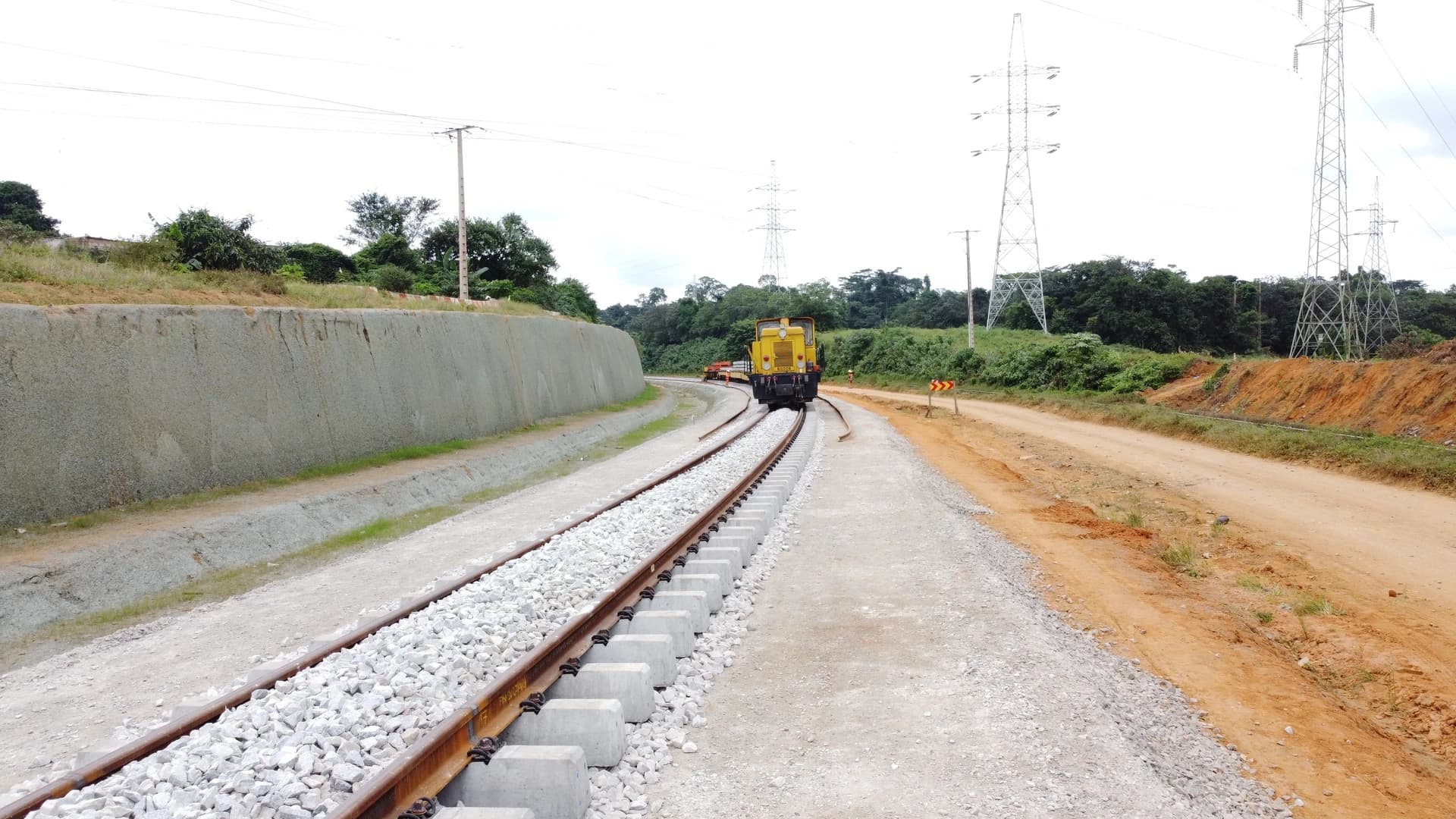 Un locotracteur jaune est visible sur une voie ferrée neuve incurvée à gauche, bordée par un mur de soutènement en béton gris à gauche et une route de terre rougeâtre à droite, avec des pylônes électriques et une végétation luxuriante en arrière-plan sous un ciel lumineux.
