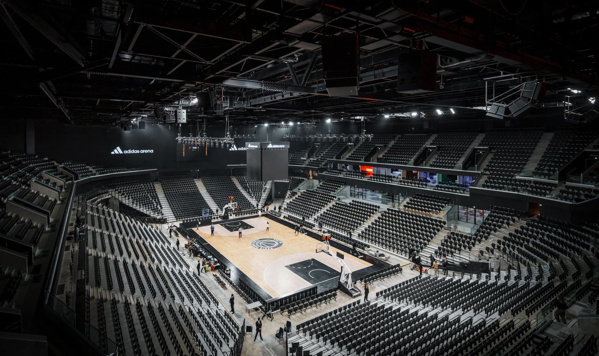 Vue d'ensemble de l'intérieur moderne de l'Adidas Arena, montrant un terrain de basketball en bois clair entouré de milliers de sièges noirs vides. Quelques personnes sont présentes sur le parquet et autour, sous un éclairage tamisé.