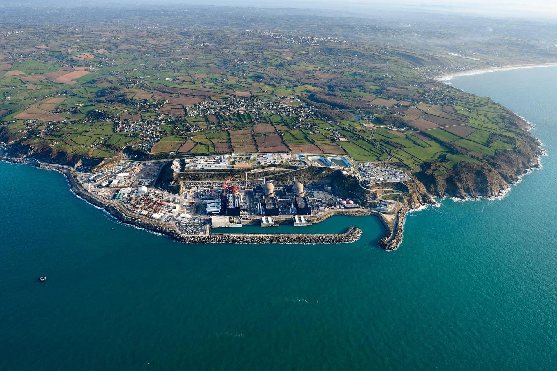 Vue aérienne de la centrale nucléaire de Flamanville sur la côte normande, montrant deux réacteurs opérationnels et un troisième en construction avec une grue rouge, entourée par la mer bleu-vert et un paysage de champs agricoles verdoyants.
