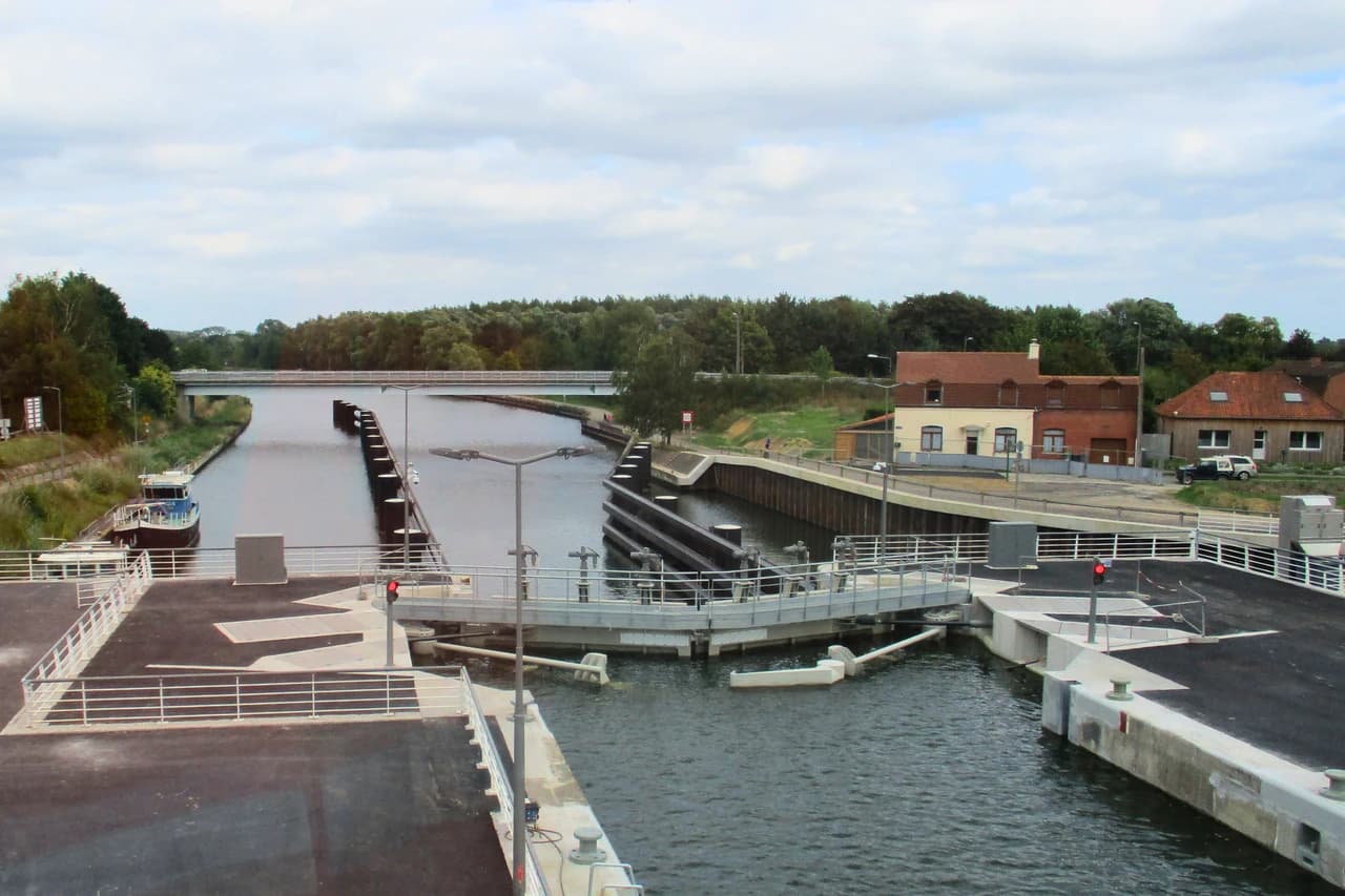 Vue aérienne d'un système d'écluse ou de pont-levis sur un canal, avec des portes partiellement ouvertes, une péniche amarrée à gauche et un pont blanc au loin. Des maisons aux toits rouges et une dense végétation bordent le canal sous un ciel nuageux.