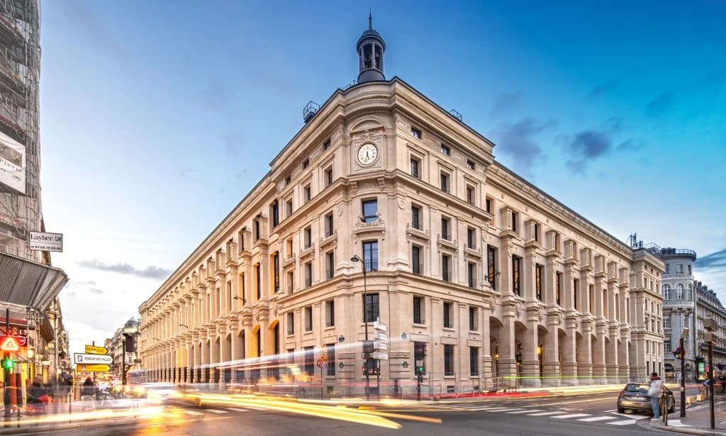Un grand bâtiment historique de style haussmannien avec une horloge sur sa façade et un dôme, situé à un coin de rue animé avec des traînées lumineuses de véhicules au crépuscule.