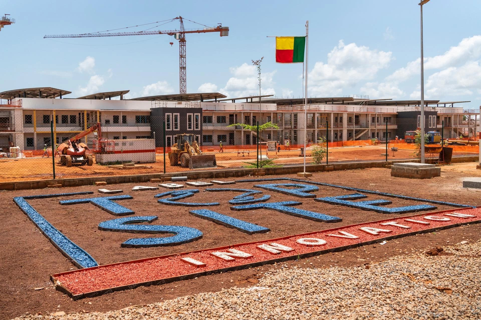 Vue d'un vaste chantier de construction ensoleillé montrant des bâtiments modernes en cours, des grues et engins, avec le drapeau du Bénin flottant. Au premier plan, des lettres bleues et le mot "INNOVATION" en rouge et blanc sont formés sur le sol de terre ocre.