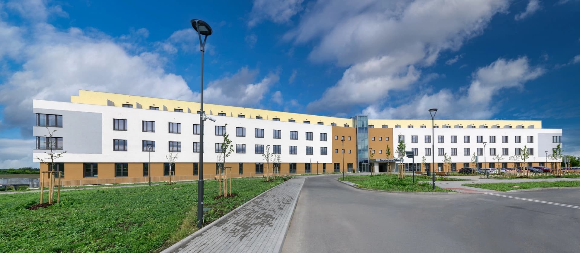 Bâtiment moderne de sanatorium incurvé aux murs blancs et bruns sous un ciel bleu nuageux avec une allée pavée.