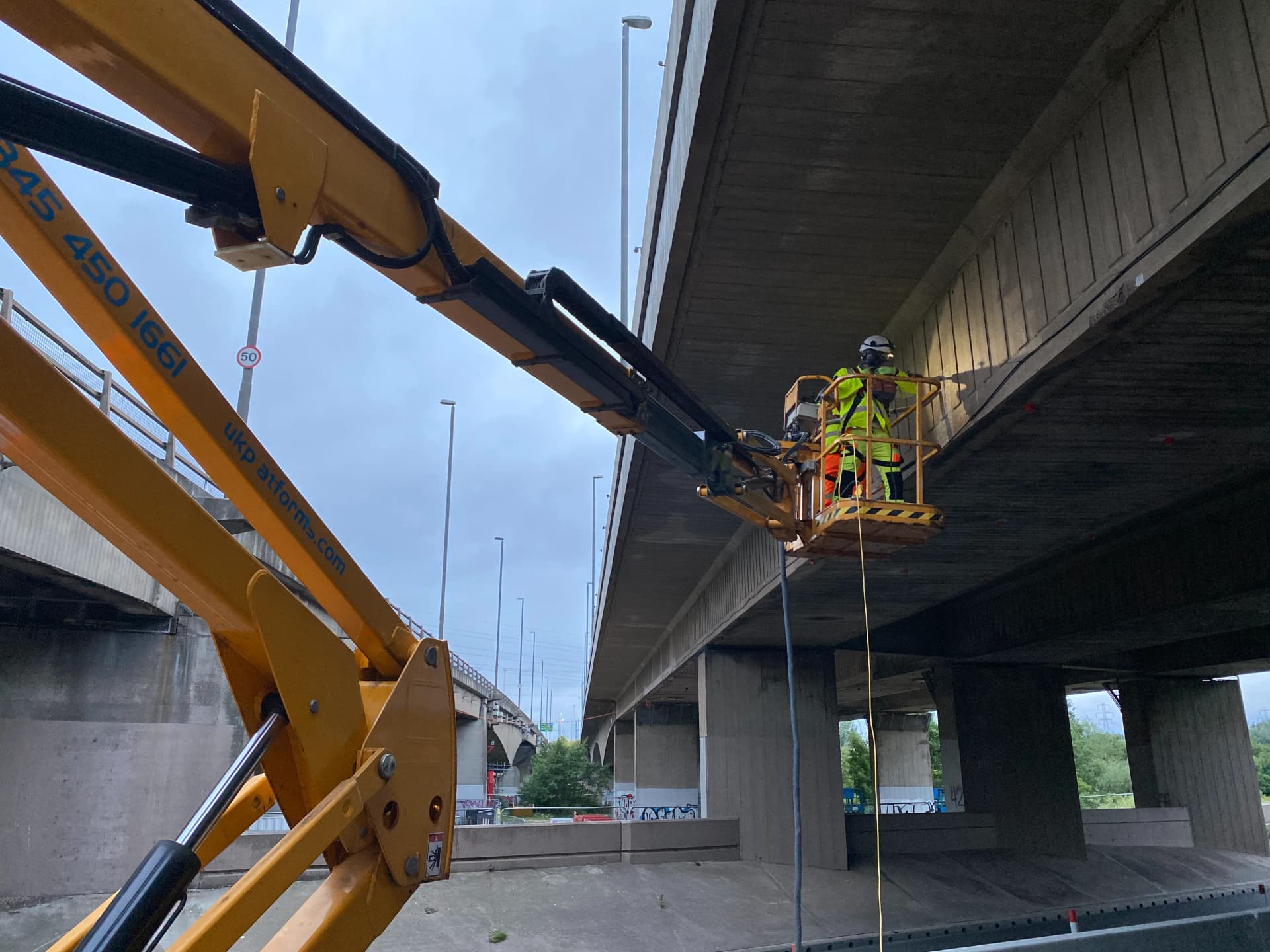 Intervention de maintenance sous un pont routier réalisée par un technicien équipé d’EPI dans une nacelle élévatrice. L’opération concerne l’inspection et la réparation de la structure en béton du tablier, illustrant des travaux spécialisés en génie civil et maintenance d’infrastructures routières.