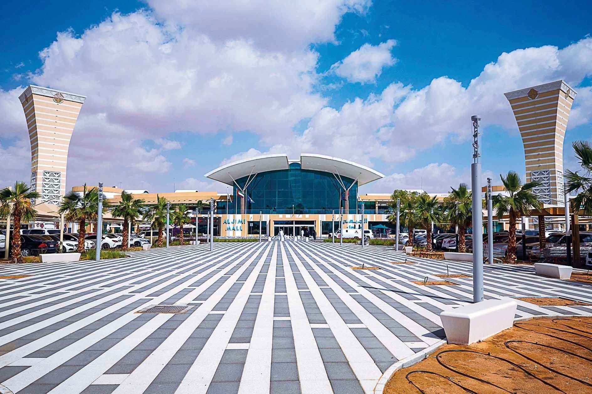 Vue extérieure de l'Al Jimi Mall avec sa place rayée, ses deux tours et son entrée vitrée sous un ciel bleu nuageux.