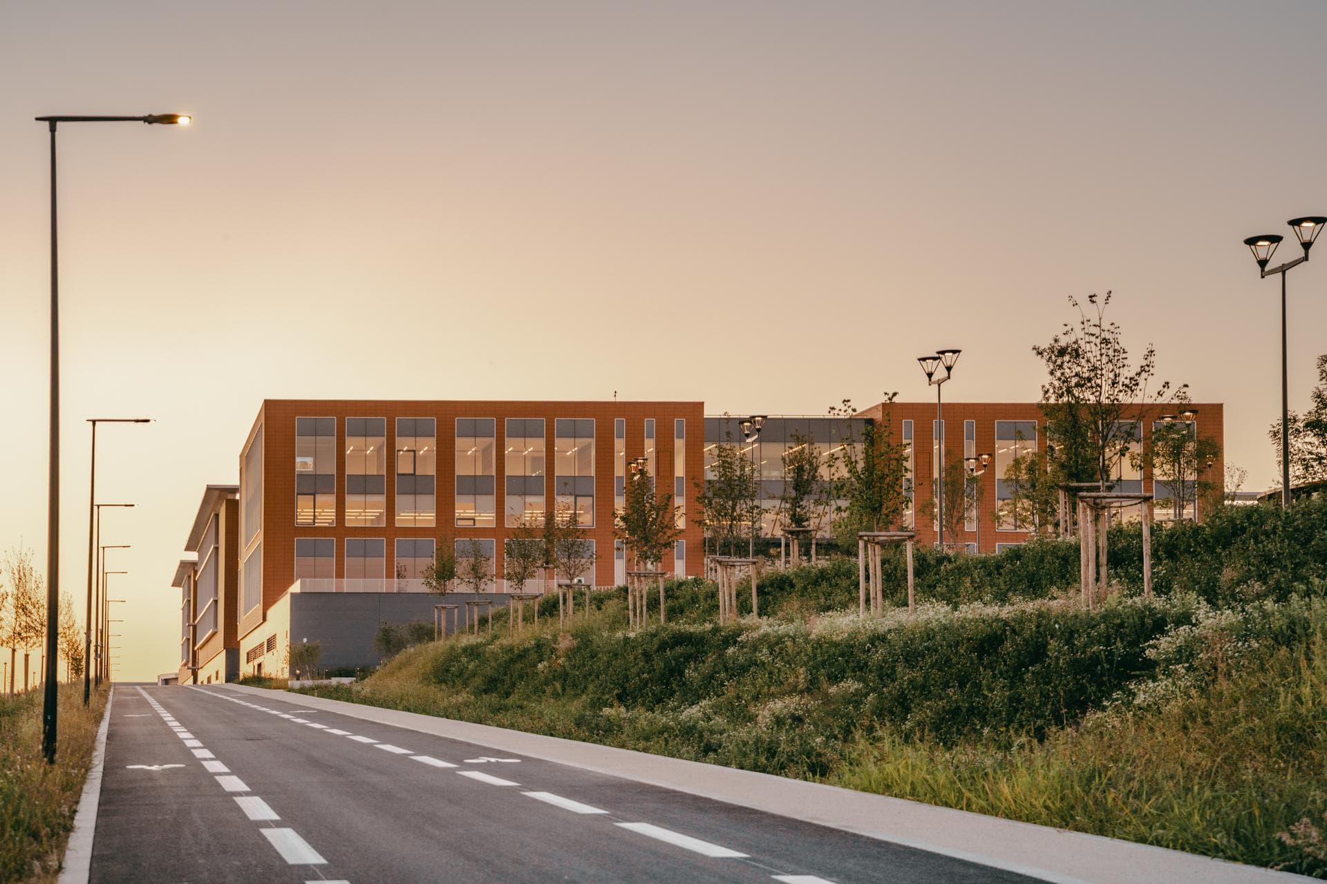 Bâtiment moderne aux façades orange et grandes fenêtres reflétant un ciel de crépuscule orangé, s'élevant derrière un talus verdoyant et une route montante éclairée par des lampadaires.