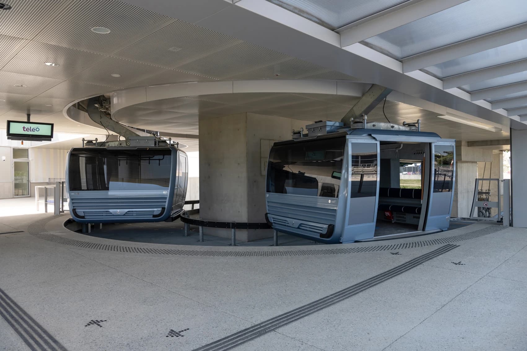 Deux cabines de téléphérique modernes, gris clair, sont garées dans une station lumineuse avec des piliers en béton et un plafond perforé. La cabine de droite a ses portes ouvertes, et un panneau vert lumineux affichant "téléo" est visible à gauche.