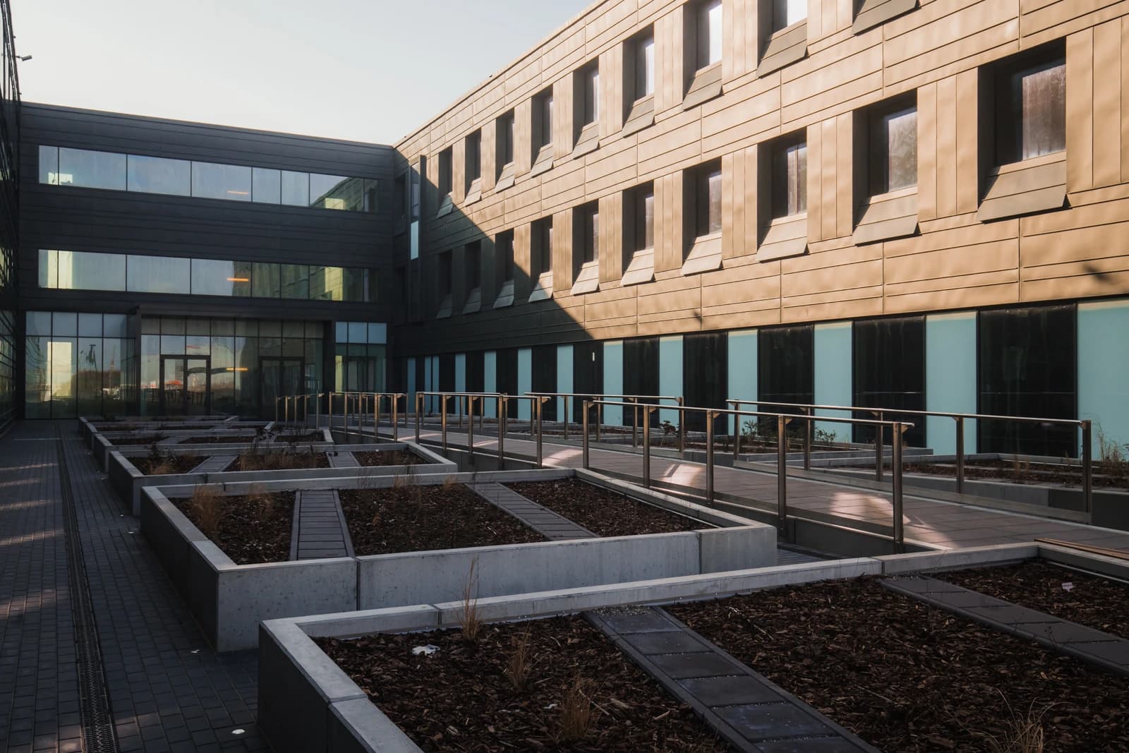 Un atrium architectural moderne avec deux façades distinctes: l'une en verre sombre et l'autre en panneaux cuivrés, bordant des jardinières rectangulaires en béton remplies de paillis et des allées pavées. Le soleil éclaire les balustrades métalliques et projette de longues ombres sur l'ensemble.