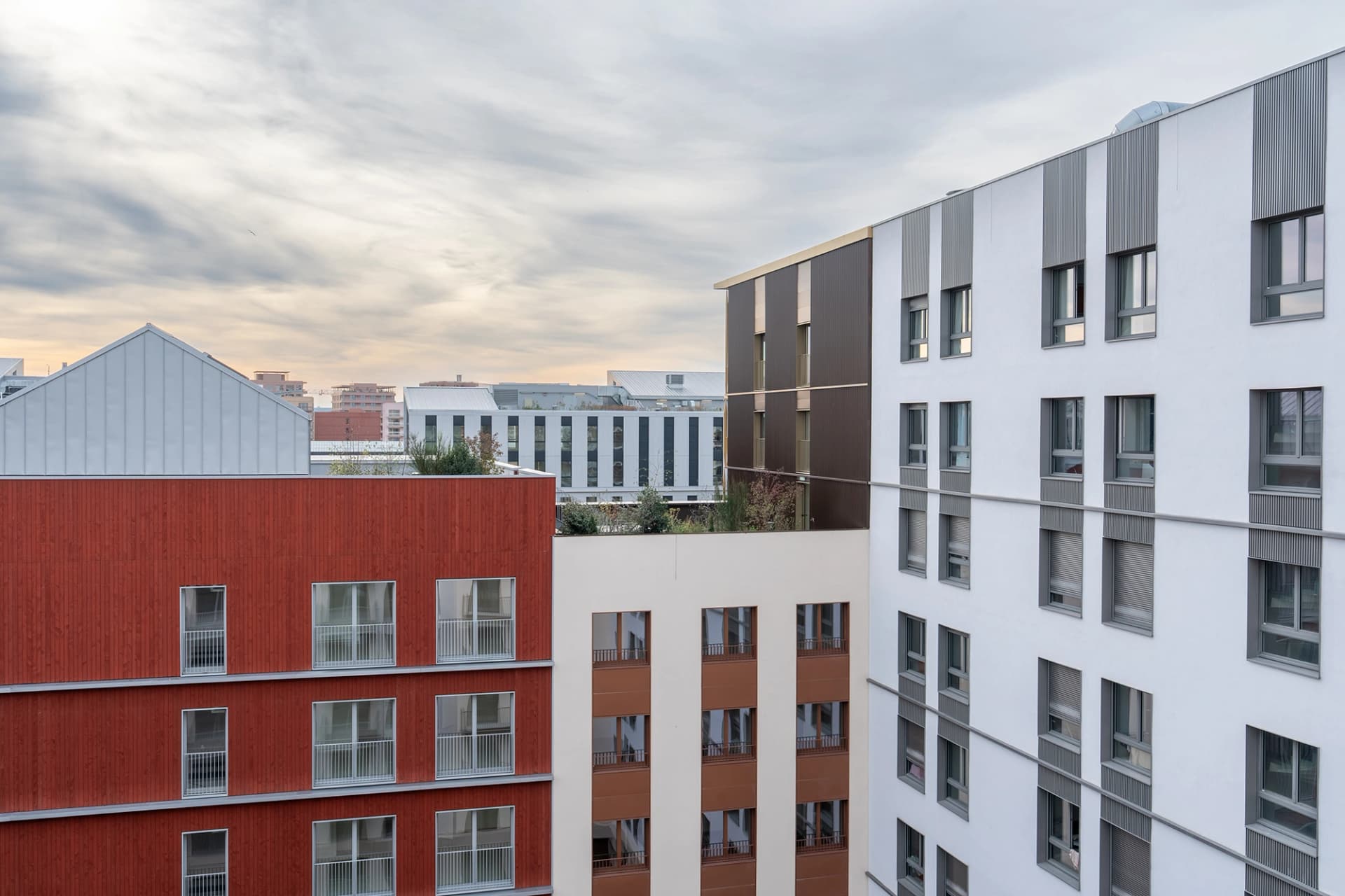 Plusieurs immeubles d'habitation modernes aux façades rouge brique, blanche et beige clair avec des fenêtres et des balcons, se dressent sous un ciel nuageux.
