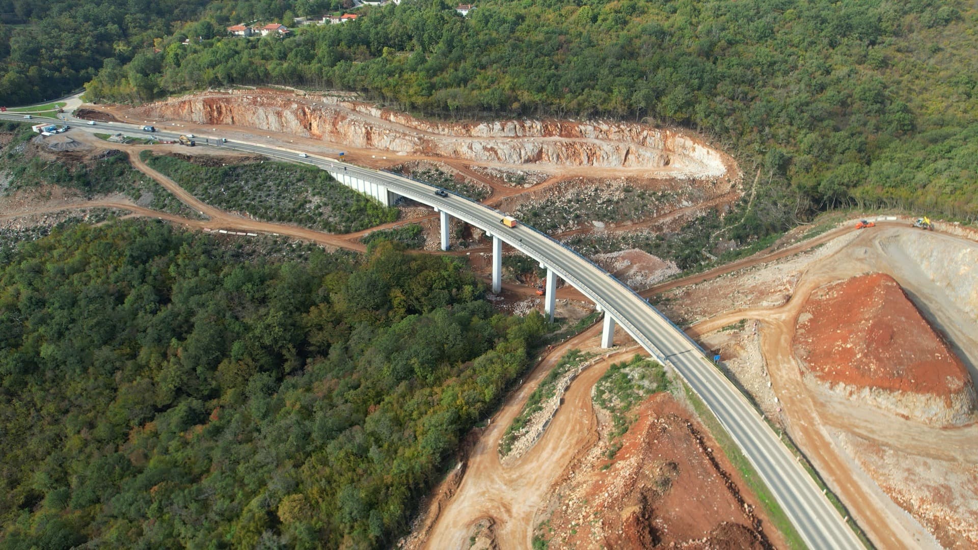 Vue aérienne d'un viaduc d'autoroute moderne en béton traversant une dense forêt verte, bordé de vastes excavations de terre rouge-orangée et de parois rocheuses, indiquant une construction récente ou en cours.