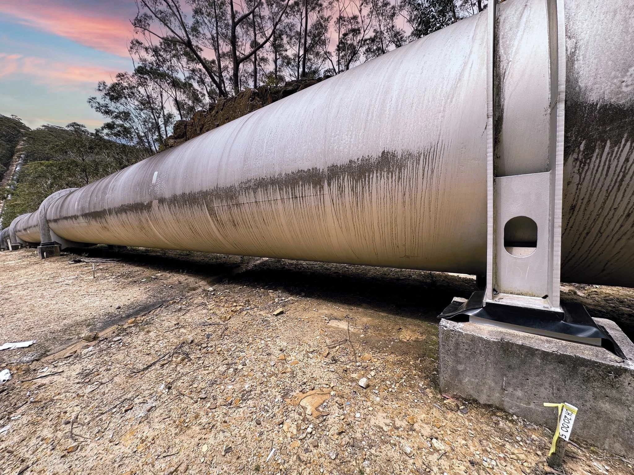 Un immense pipeline en métal gris patiné et strié traverse un terrain sec et rocailleux, soutenu par des blocs de béton. En arrière-plan, des arbres élancés se dressent sous un ciel de coucher de soleil aux teintes vives de rose, d'orange et de bleu.