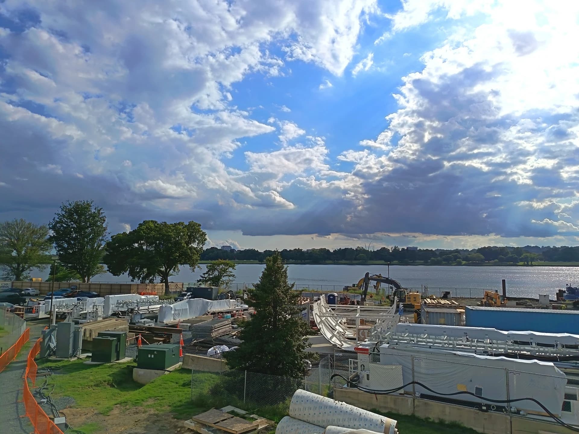 Un chantier de construction s'étend au bord de l'eau, avec des équipements et des matériaux bâchés, sous un ciel bleu éclatant parsemé de nuages blancs et gris où filtrent des rayons de soleil.