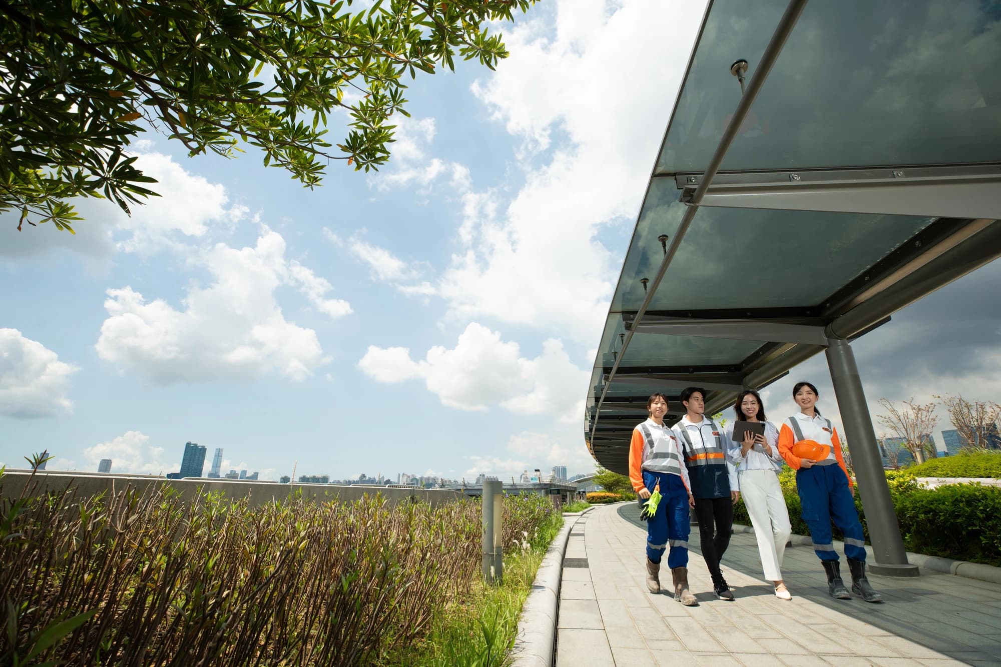 Quatre jeunes adultes, trois en tenue de travail orange et bleue et une en tenue décontractée blanche, marchent le long d'un chemin extérieur moderne sous un auvent de verre. Ils tiennent une tablette et un casque de sécurité, avec une silhouette de ville et de la verdure sous un ciel bleu nuageux en arrière-plan.