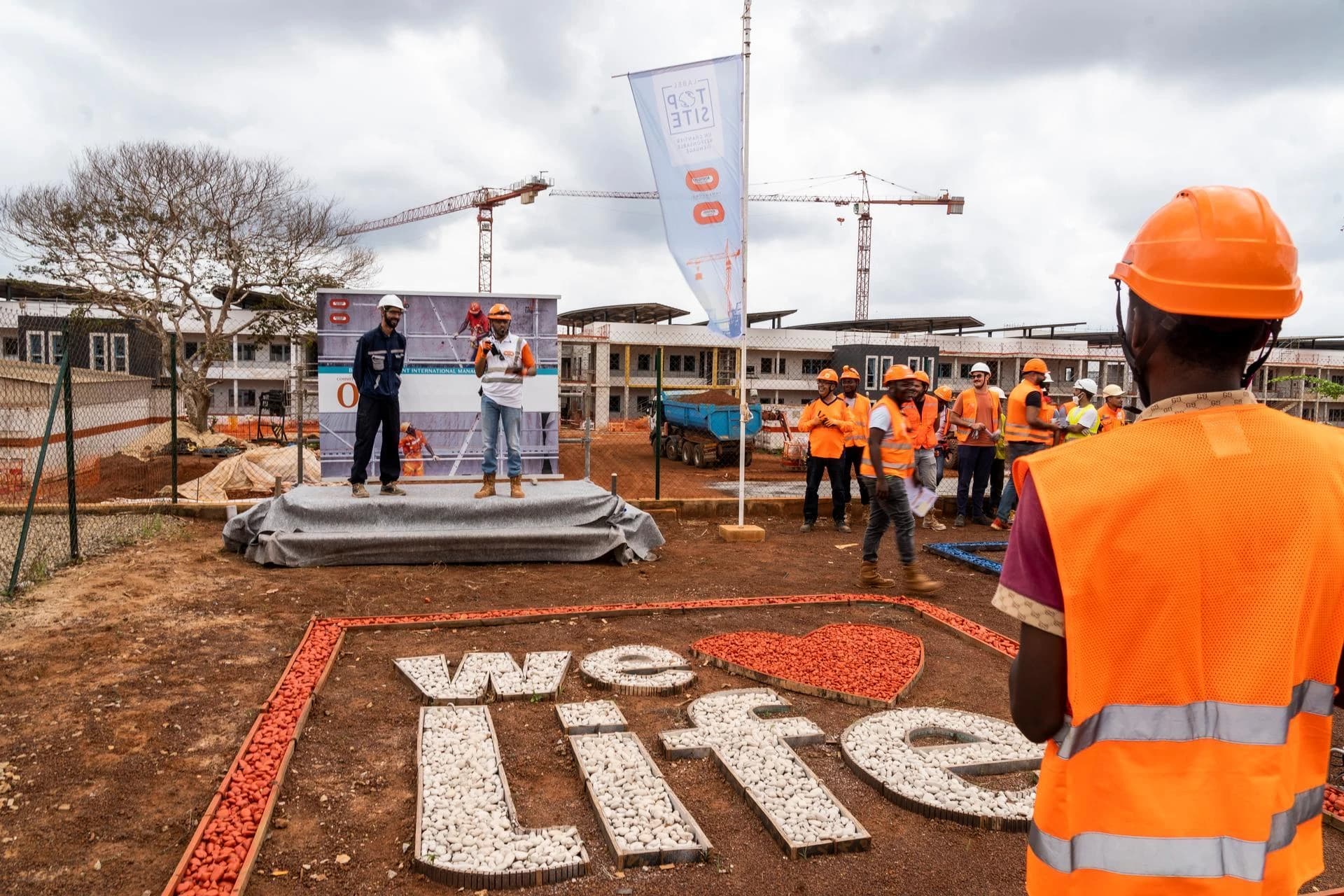 Des ouvriers du bâtiment en gilets et casques de sécurité orange sont rassemblés sur un chantier en terre battue, écoutant une personne parler depuis une estrade. Des grues sont visibles à l'arrière-plan, et au premier plan, "we ❤️ life" est écrit avec des pierres sur le sol.