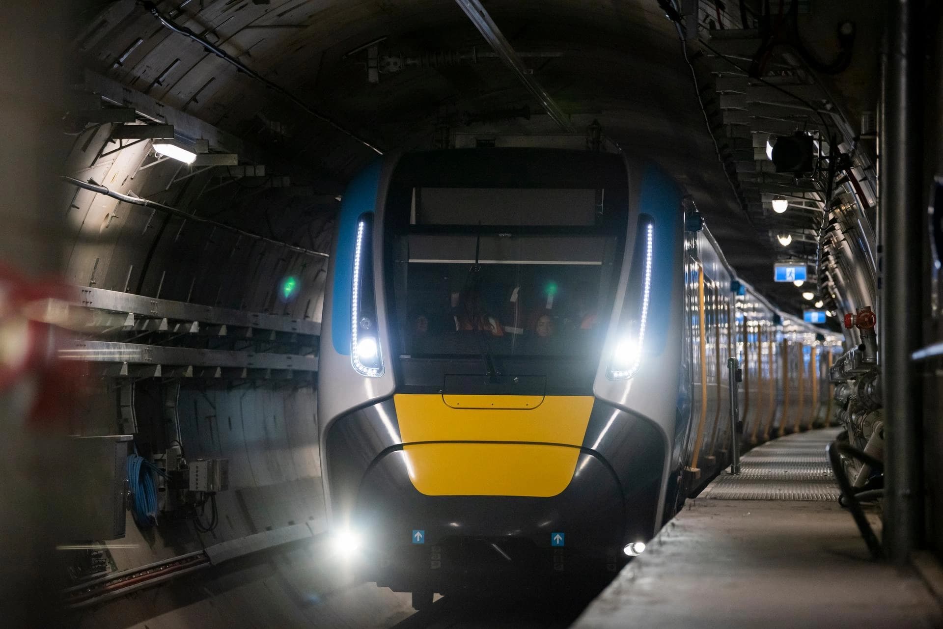 Un train moderne aux phares vifs, de couleur bleue, grise et jaune, approche d'une gare souterraine sombre. Quelques personnes sont visibles à travers les fenêtres du conducteur, tandis que le tunnel est éclairé par des lumières industrielles et des signaux verts.