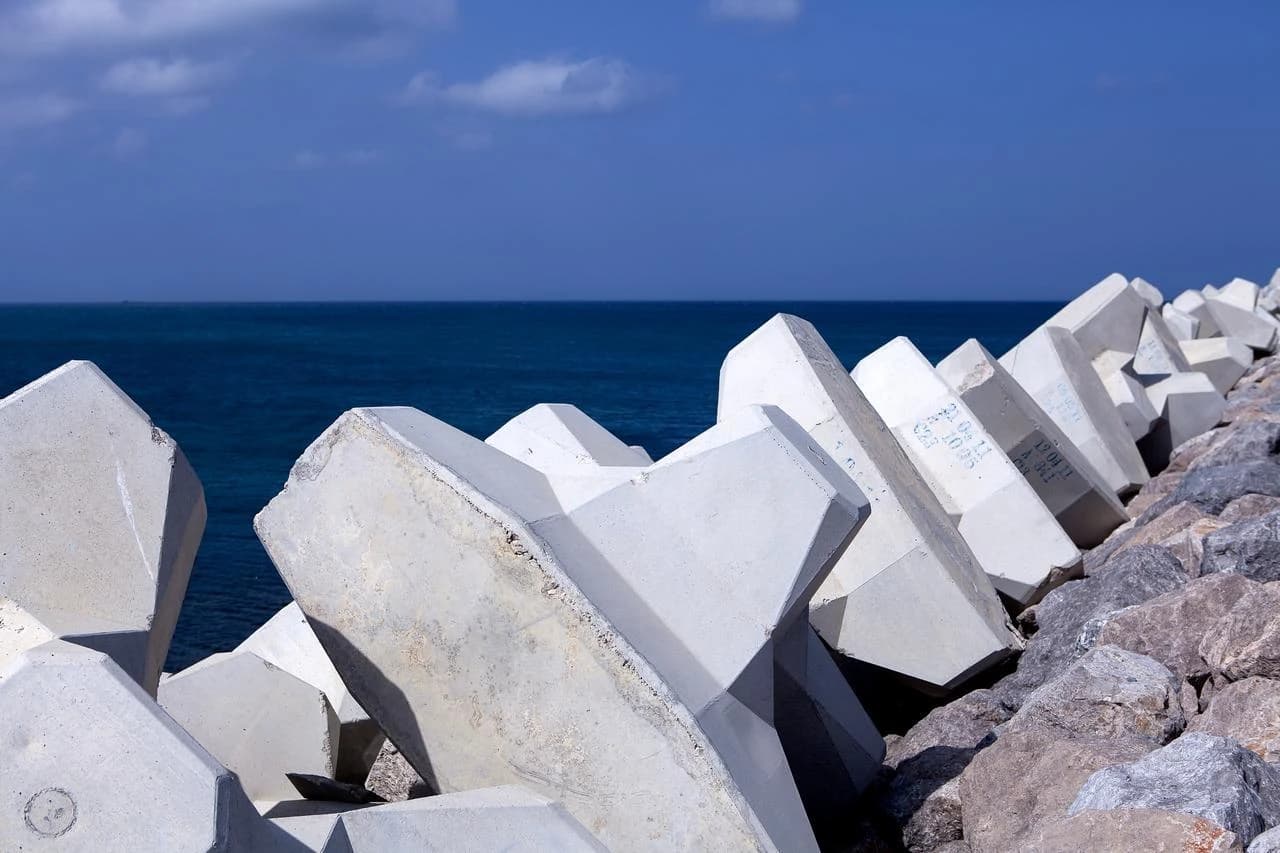 Une série de tétrapodes massifs en béton gris clair et de roches forment une digue le long d'une côte, avec l'océan d'un bleu profond sous un ciel clair en arrière-plan.