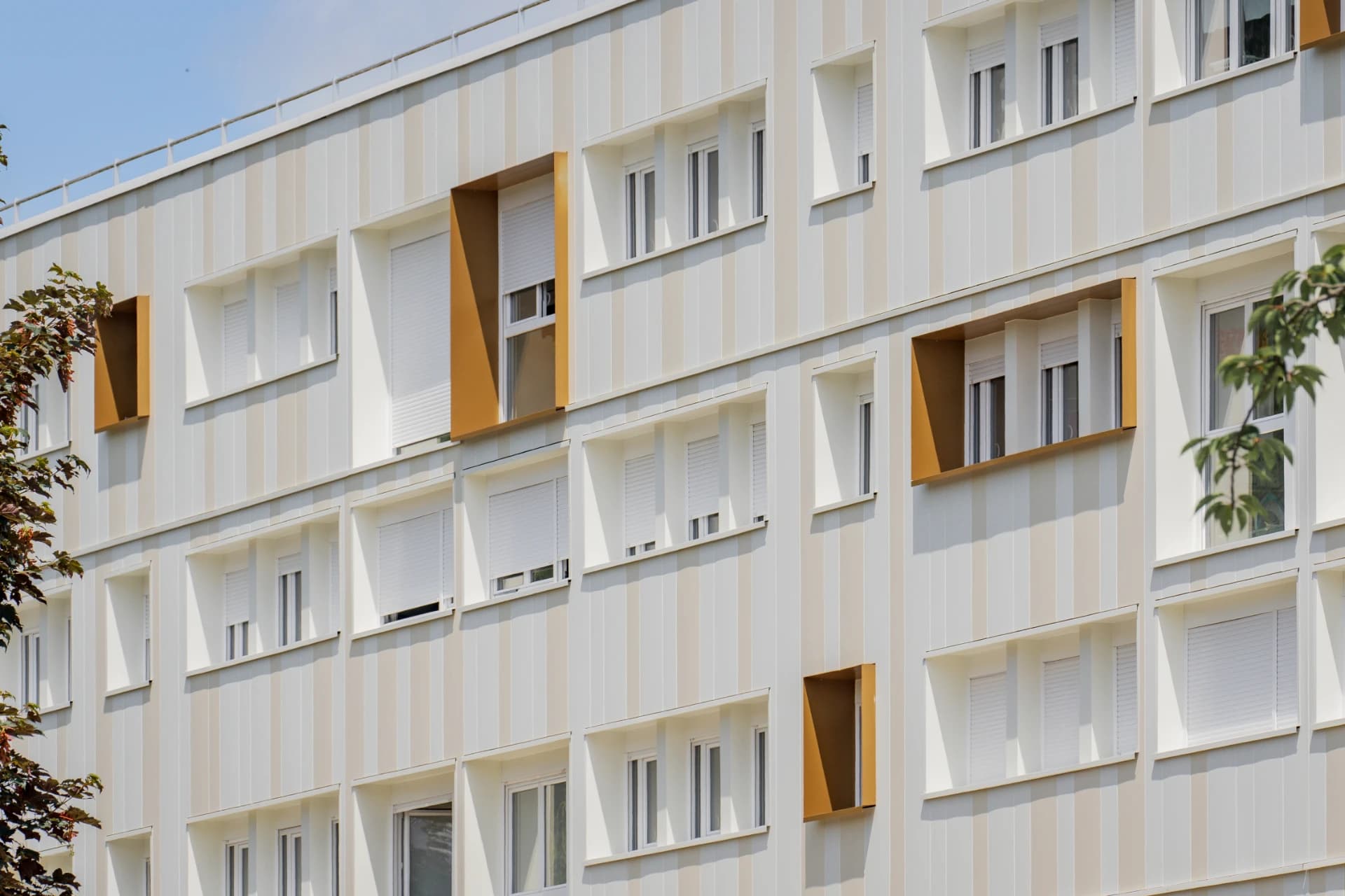 Façade moderne d'un bâtiment avec des bandes verticales alternées gris clair et beige, ponctuée de fenêtres à stores blancs et d'encadrements profonds de couleur orange-brun, sous un ciel bleu.