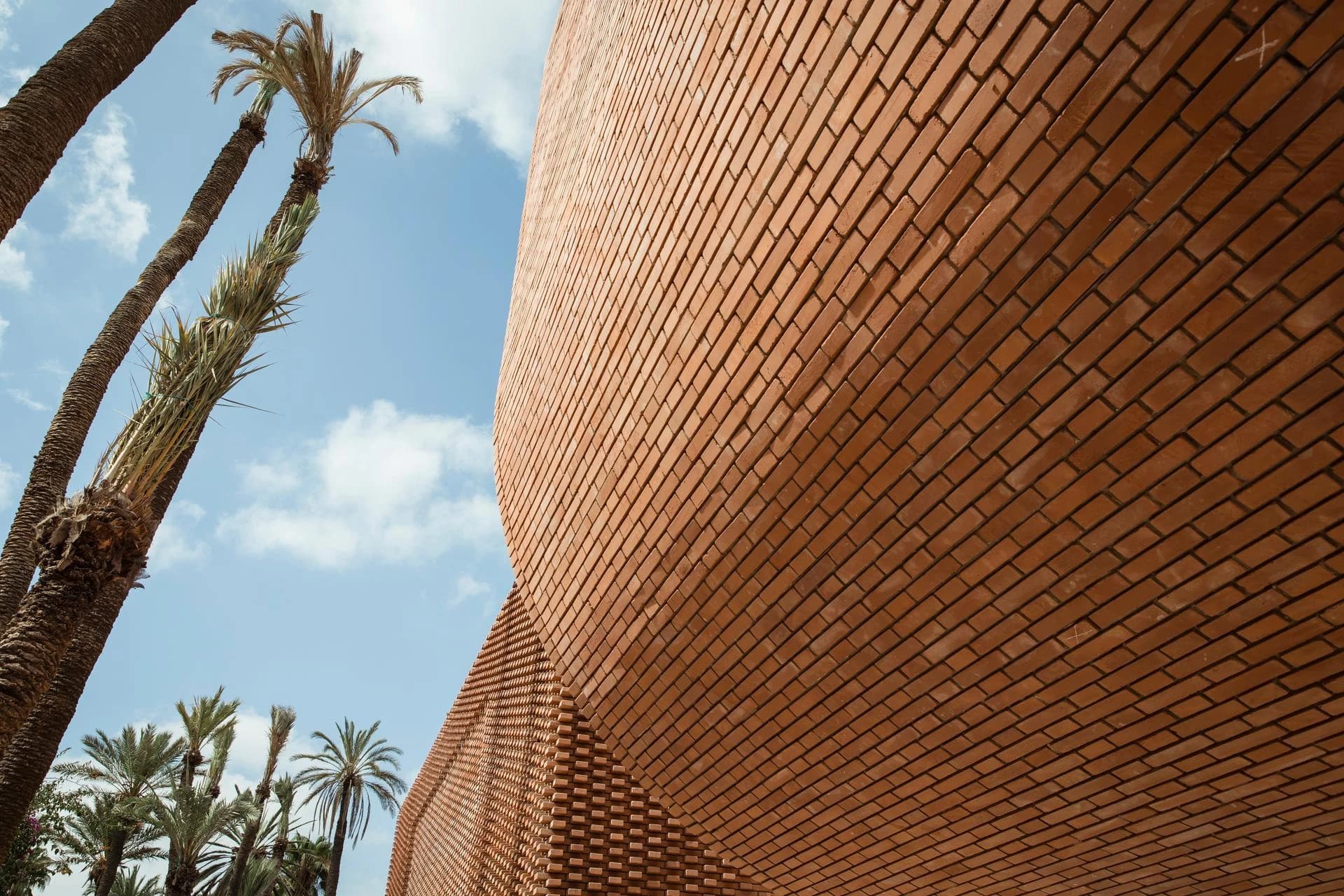 Une paroi incurvée d'un bâtiment moderne en briques orange-rouge, avec une texture en relief à sa base, se dresse à côté de hauts palmiers sous un ciel bleu parsemé de nuages blancs.