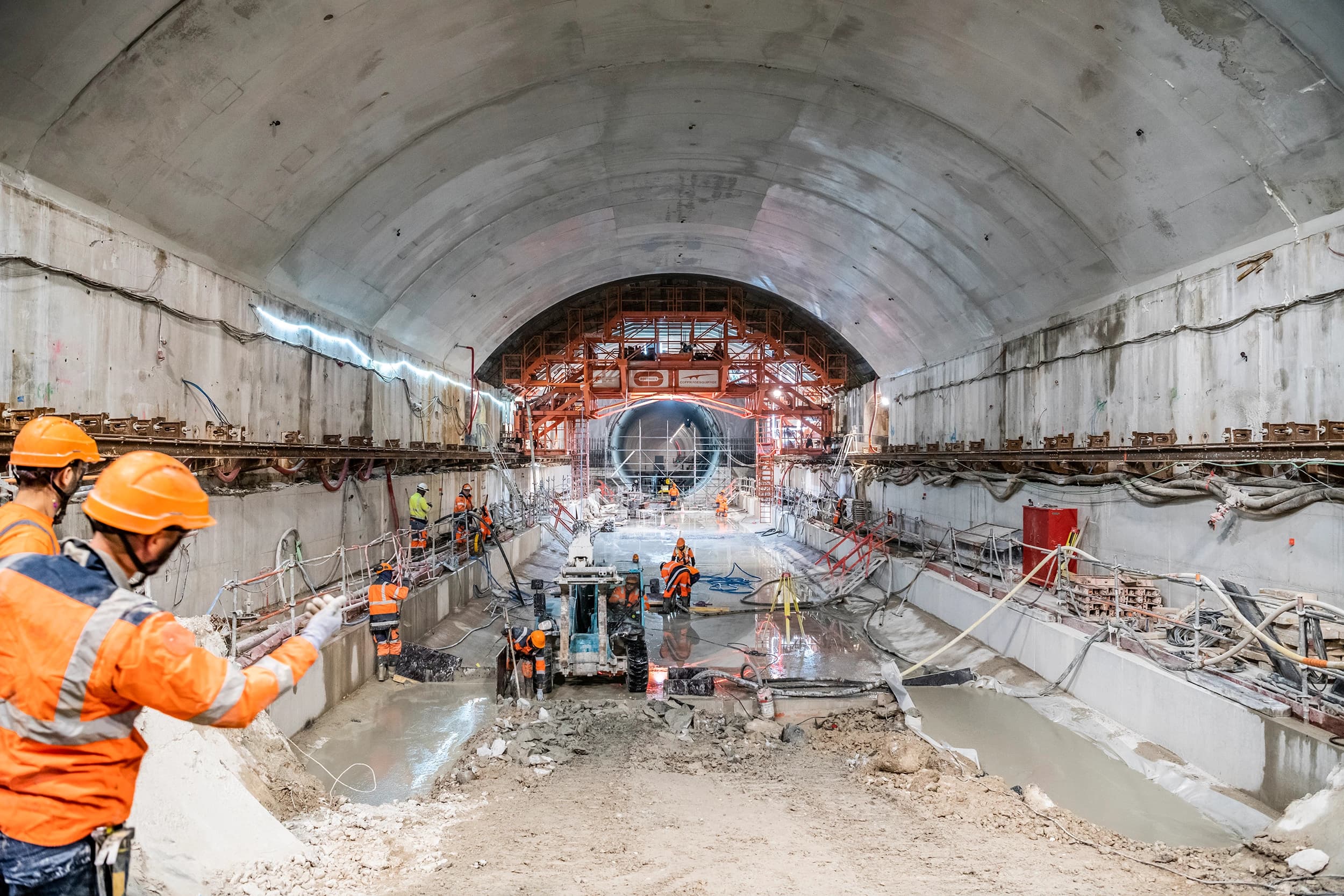 Des compagnons en tenue de sécurité orange s'affairent dans un grand tunnel de béton en construction, où le sol est humide et divers équipements sont dispersés. Au fond, une imposante structure d'échafaudage orange indique la progression des travaux, potentiellement pour le revêtement du tunnel ou une nouvelle section.
