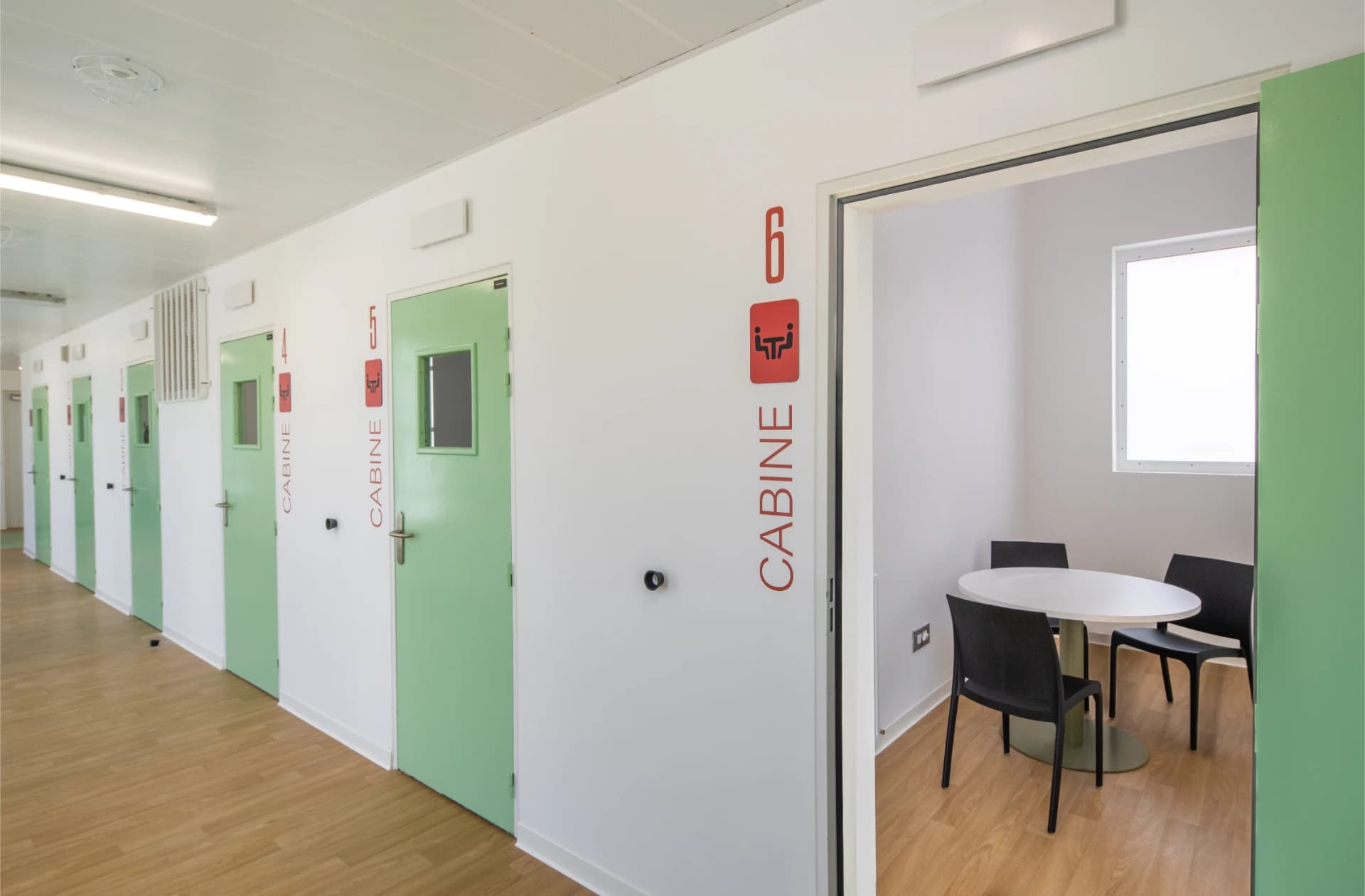 Un couloir lumineux avec un sol en bois clair présente une série de portes vert pâle, chacune marquée "CABINE" avec un numéro et une icône de réunion en rouge. La sixième cabine, légèrement ouverte, révèle un petit espace avec une table ovale blanche et trois chaises noires.