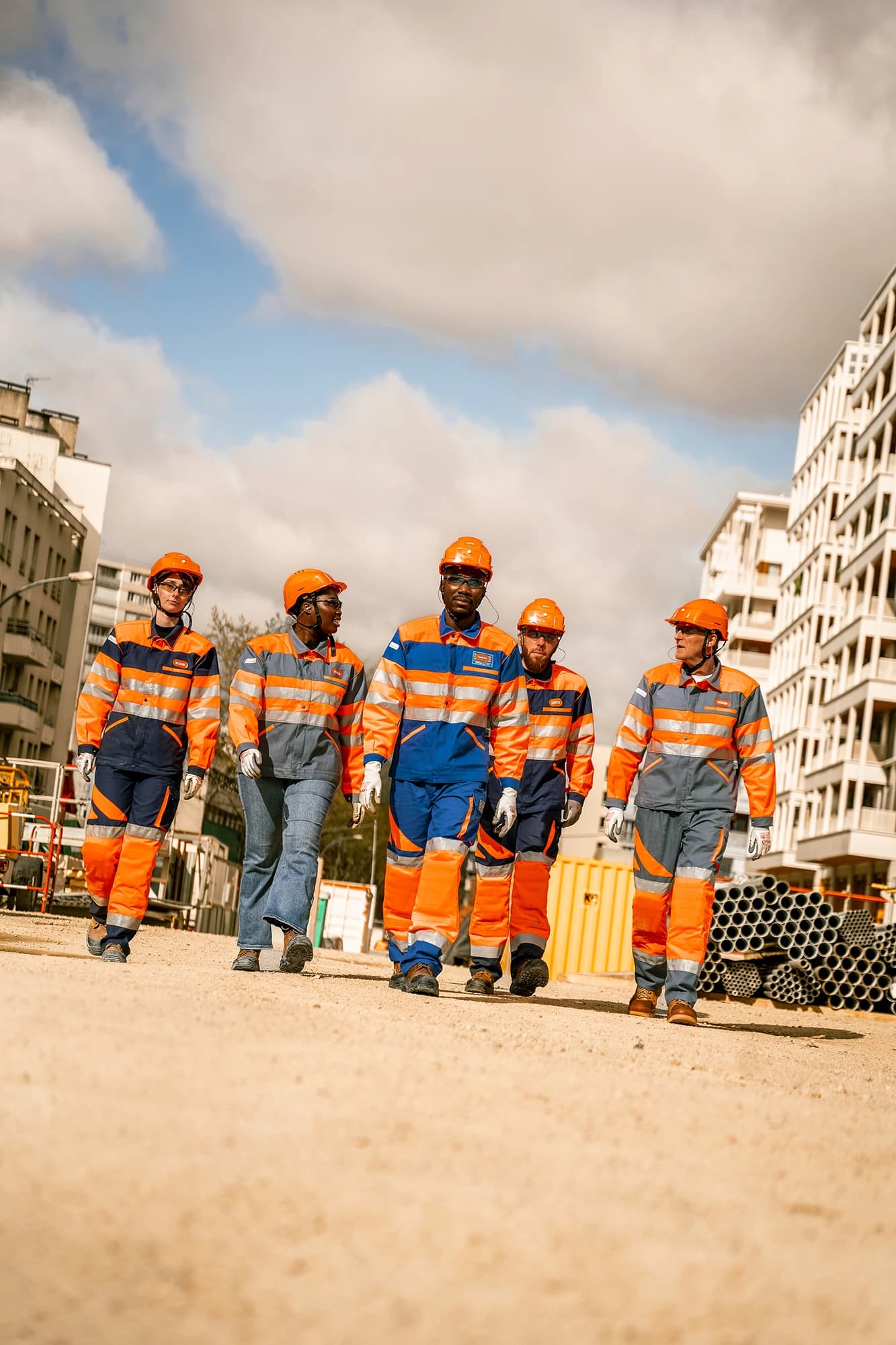Une équipe de cinq ouvriers, portant des casques de sécurité orange et des uniformes haute visibilité orange, bleus et gris, marche sur un sol terreux. À l'arrière-plan, des bâtiments urbains et des matériaux de construction sont visibles sous un ciel partiellement nuageux.