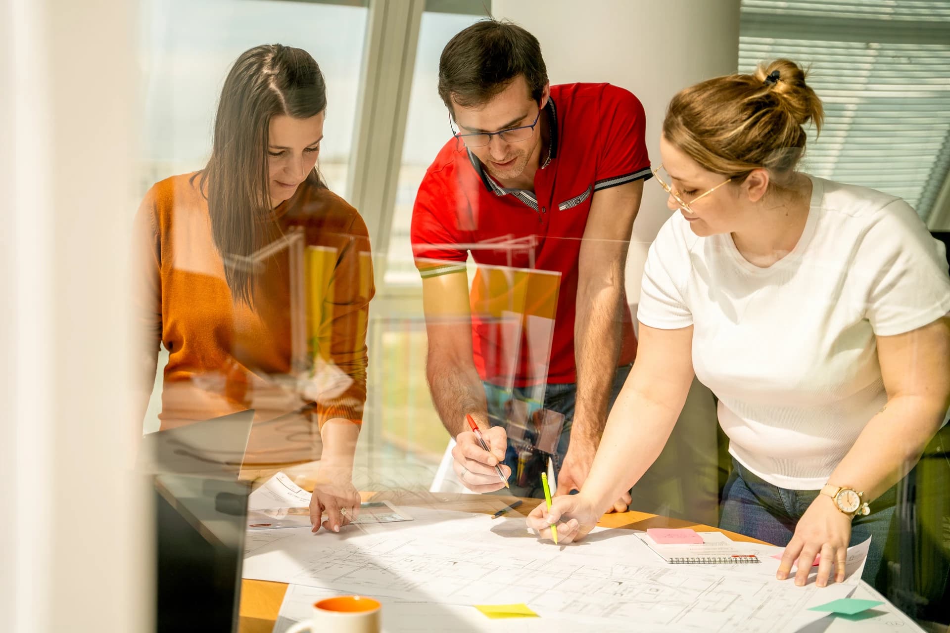 Trois collègues, deux femmes et un homme, se penchent sur une table, examinant des plans architecturaux et prenant des notes avec des stylos dans un bureau lumineux.
