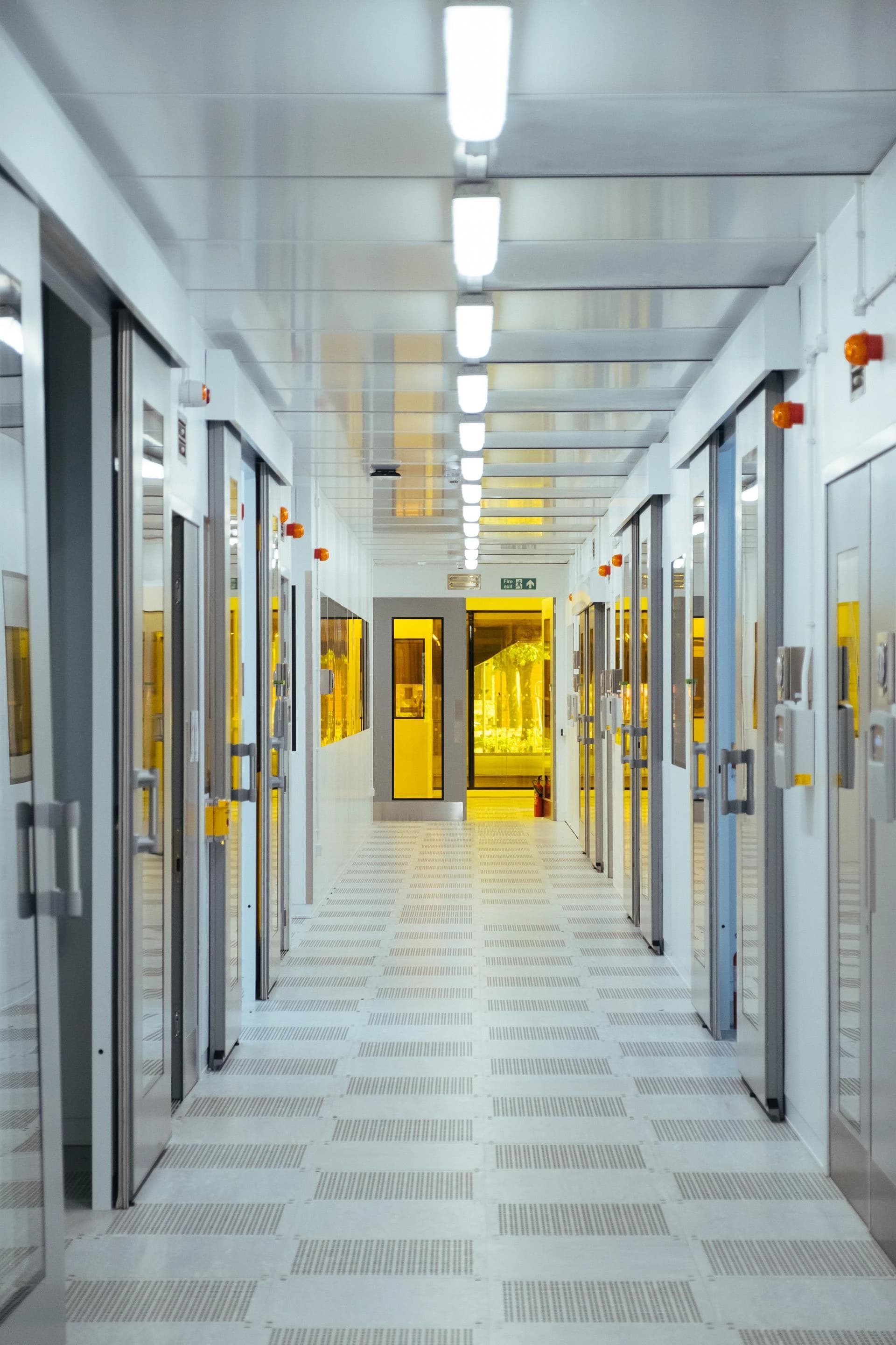 Un long couloir stérile et lumineux dans une installation de haute technologie est bordé de portes blanches et réfléchissantes de chaque côté. Une lumière jaune vif émane des pièces, contrastant avec l'éclairage blanc froid du plafond et le sol gris clair texturé.