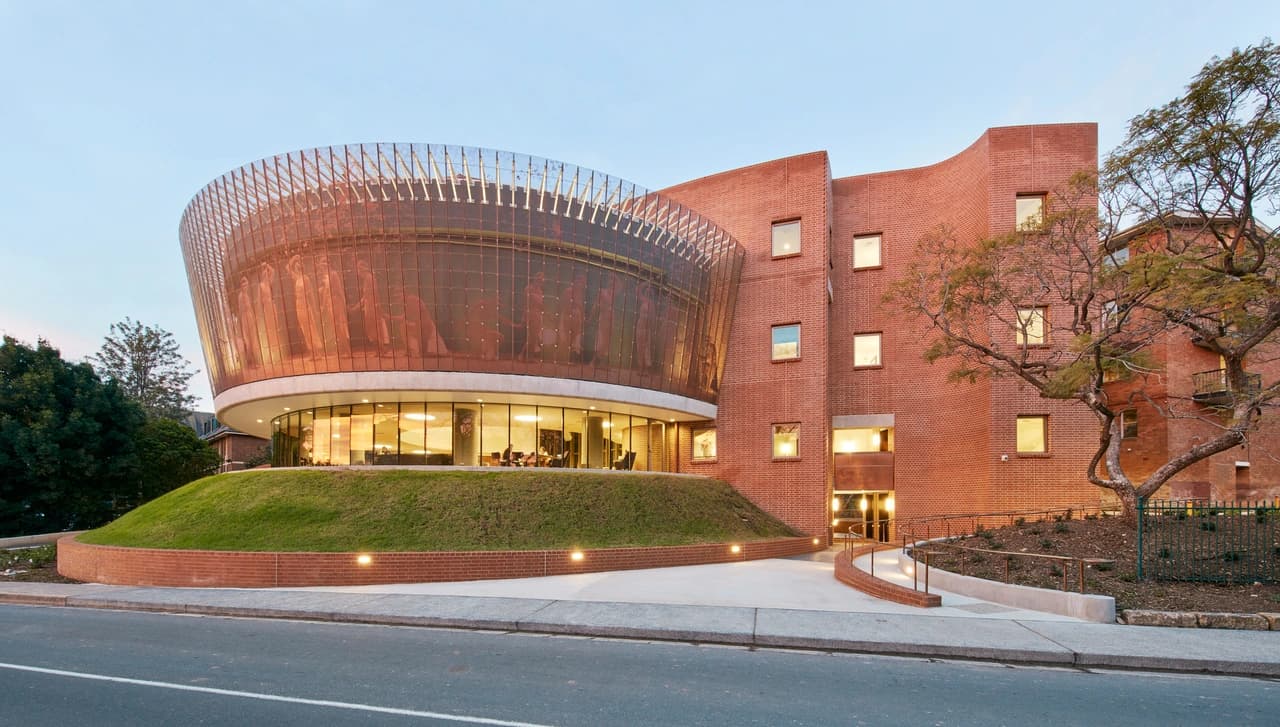Façade architecturale complexe combinant des murs en briques rouges et une structure cylindrique vitrée enveloppée d'un voile métallique perforé.