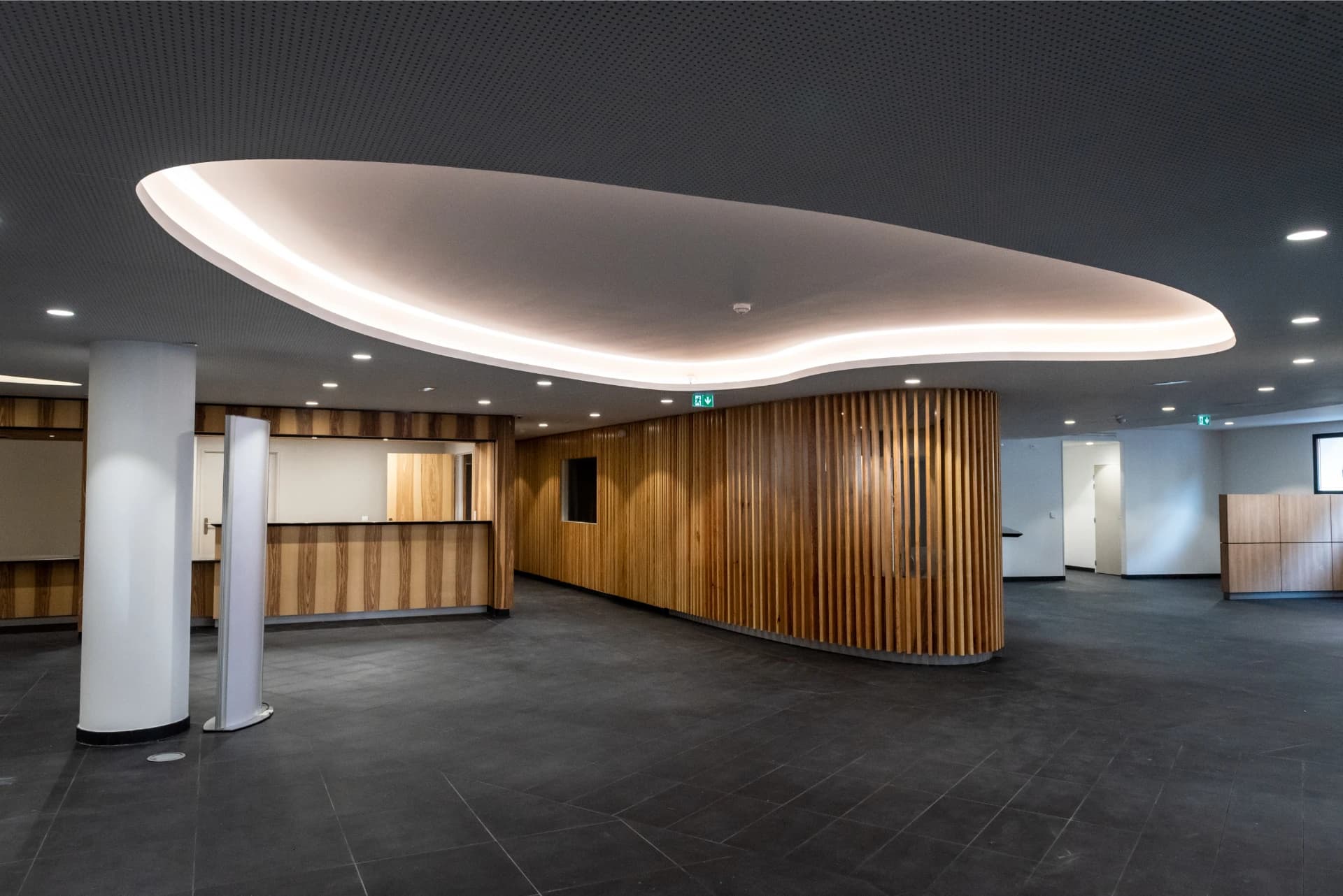 Un hall moderne et vide avec un plafond gris foncé perforé et un panneau lumineux incurvé blanc, au-dessus de murs recouverts de lattes de bois verticales chaudes et d'un sol en carrelage gris foncé. Un comptoir de réception en bois, un pilier blanc et un roll-up banner sont également visibles.