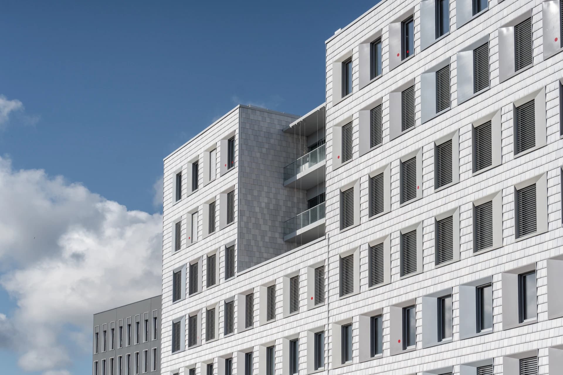 Façade moderne d'un grand immeuble blanc aux tuiles imbriquées, avec des rangées de fenêtres aux volets gris (certains ouverts) et de petits points rouges, ainsi que des balcons en verre. Un ciel bleu clair parsemé de nuages est visible à l'arrière-plan.