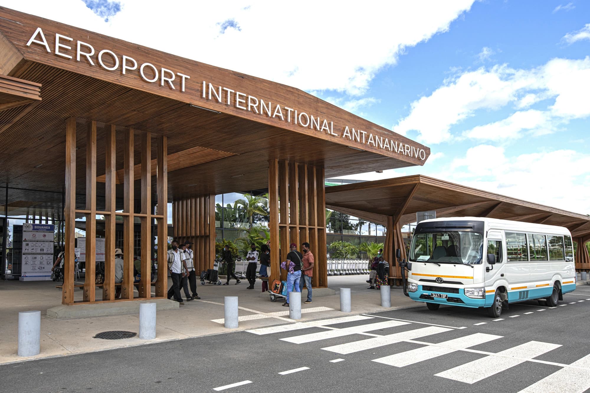 Entrée moderne de l'aéroport international d'Antananarivo avec une vaste marquise en bois marron portant son nom en lettres blanches, sous un ciel bleu nuageux. Un bus navette blanc et bleu est garé près d'un passage piéton, avec plusieurs personnes circulant à l'entrée.
