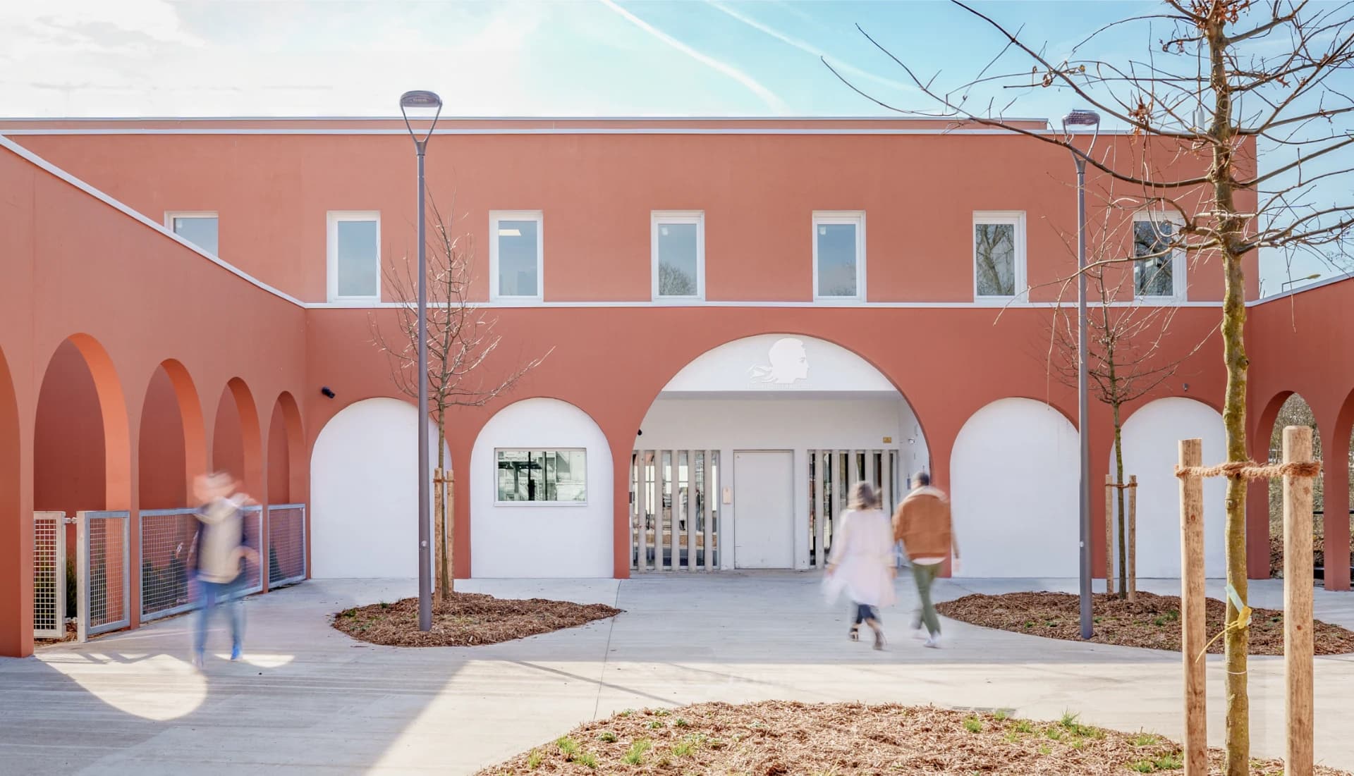 Façade moderne orange terracota d'un bâtiment public avec des arches blanches et un grand porche central orné d'une silhouette de Marianne et l'inscription "RÉPUBLIQUE FRANÇAISE", encadrant une cour ensoleillée où des personnes floues marchent.