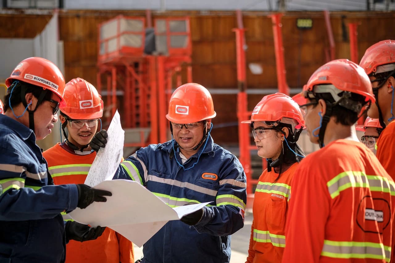 Une équipe de travailleurs de la construction portant des casques de sécurité orange, des lunettes de protection et des vêtements haute visibilité discute des plans sur un chantier extérieur. Un homme en uniforme bleu tient et indique les plans, entouré de ses collègues.