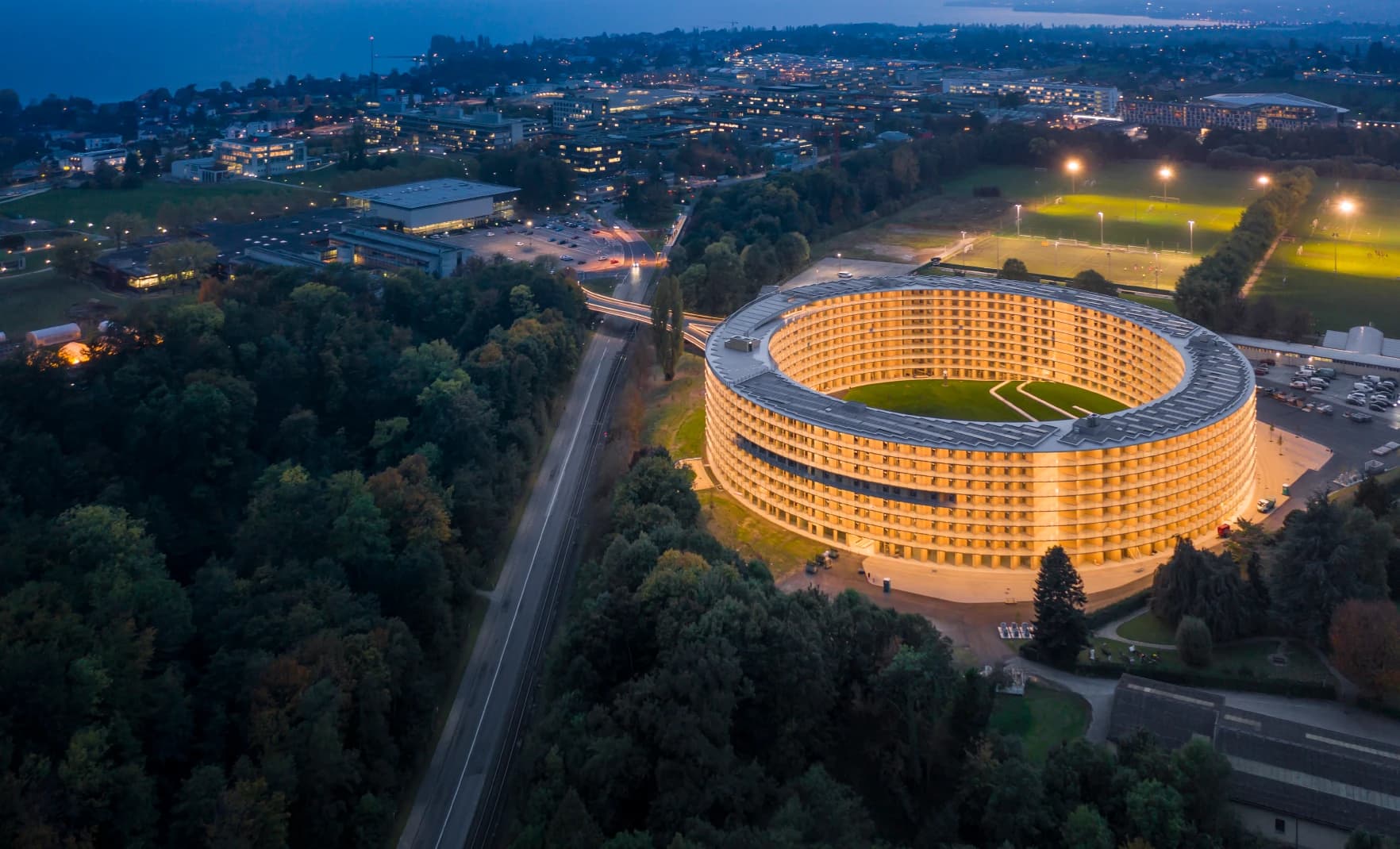 Vue aérienne nocturne d'un vaste bâtiment circulaire moderne, illuminé de lumières jaunes chaudes et doté d'une cour intérieure verte. Il est entouré de forêts sombres, de routes, de terrains de sport éclairés et d'un campus urbain lointain sous un ciel bleu foncé.