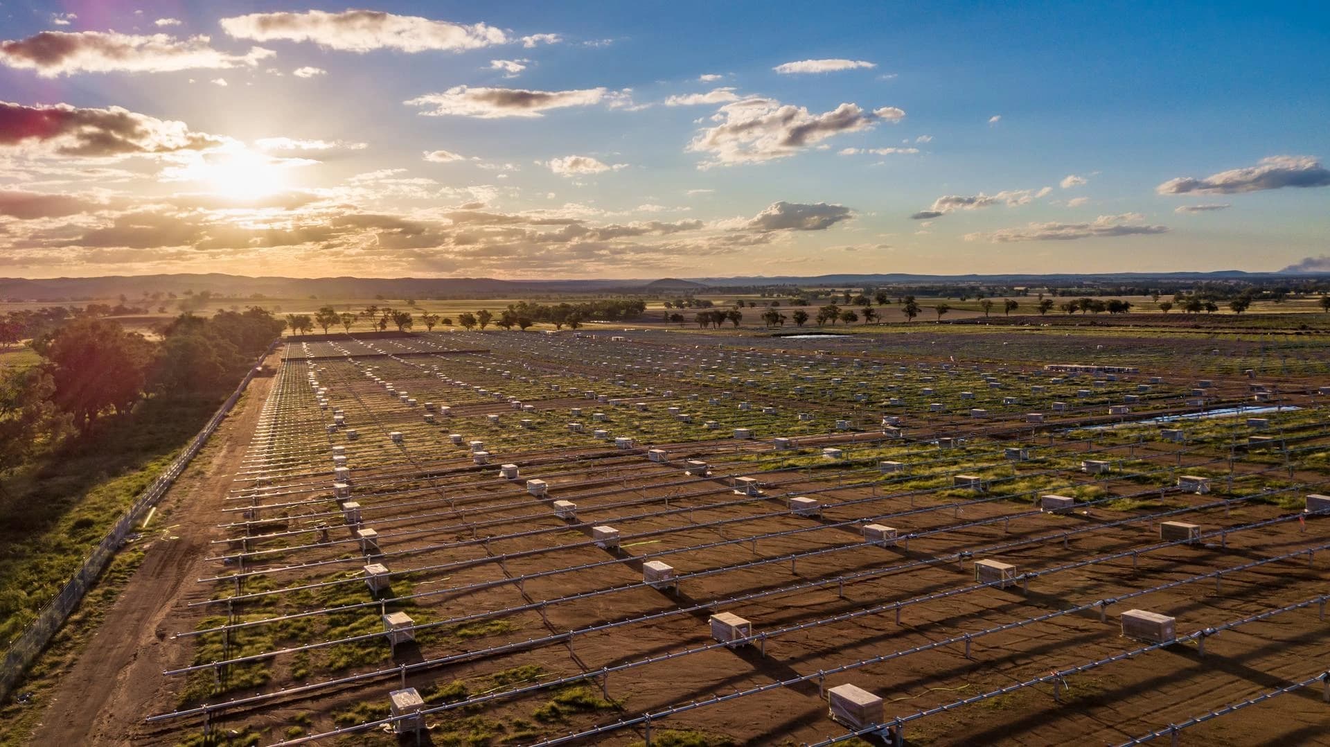 Vue aérienne d'un vaste champ de terre et d'herbe où sont installées des rangées de structures métalliques et des boîtes d'équipement pour un parc solaire. Le soleil couchant illumine l'horizon gauche d'une lumière dorée et orange, créant de longues ombres sous un ciel bleu et nuageux au-dessus d'un paysage vallonné.