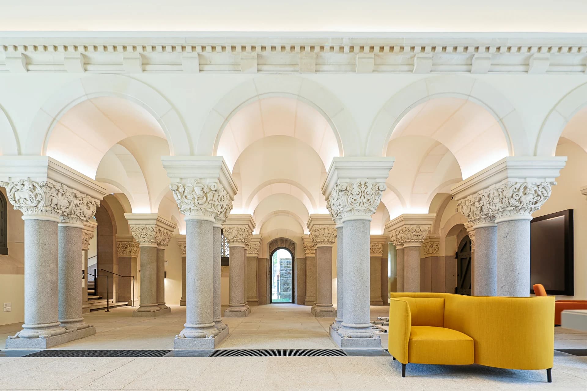 Un élégant hall présente des rangées de colonnes en pierre grise avec des chapiteaux blancs richement sculptés, soutenant des arches blanches légèrement éclairées menant à une porte au fond. Un canapé moderne incurvé jaune moutarde se détache sur le sol en pierre claire au premier plan.