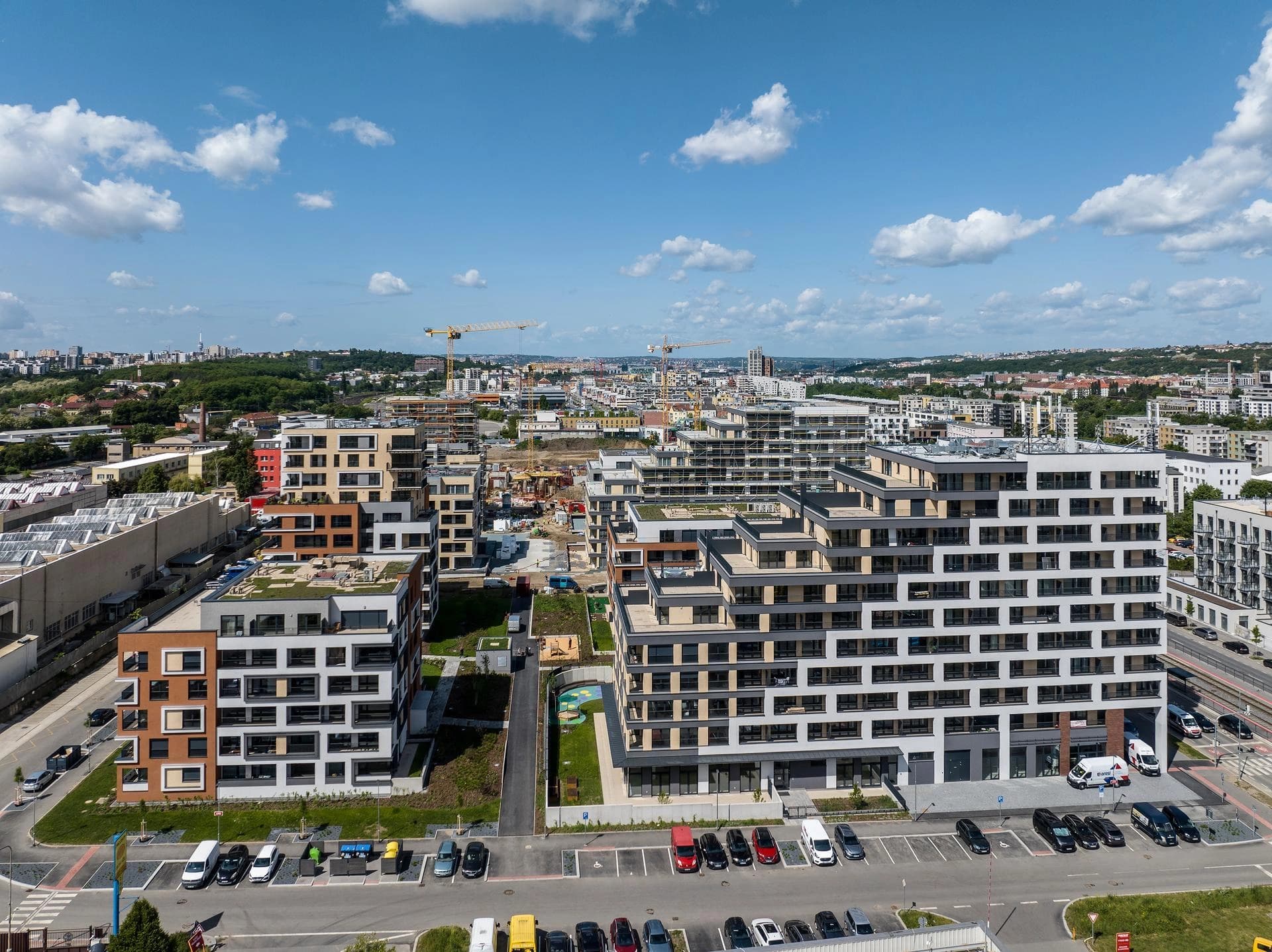 Vue aérienne d'un complexe résidentiel moderne comprenant plusieurs immeubles aux façades blanches, grises et terracotta avec des toits verts. Le paysage urbain en arrière-plan montre des chantiers avec des grues, des parkings et d'autres bâtiments, sous un ciel bleu nuageux.