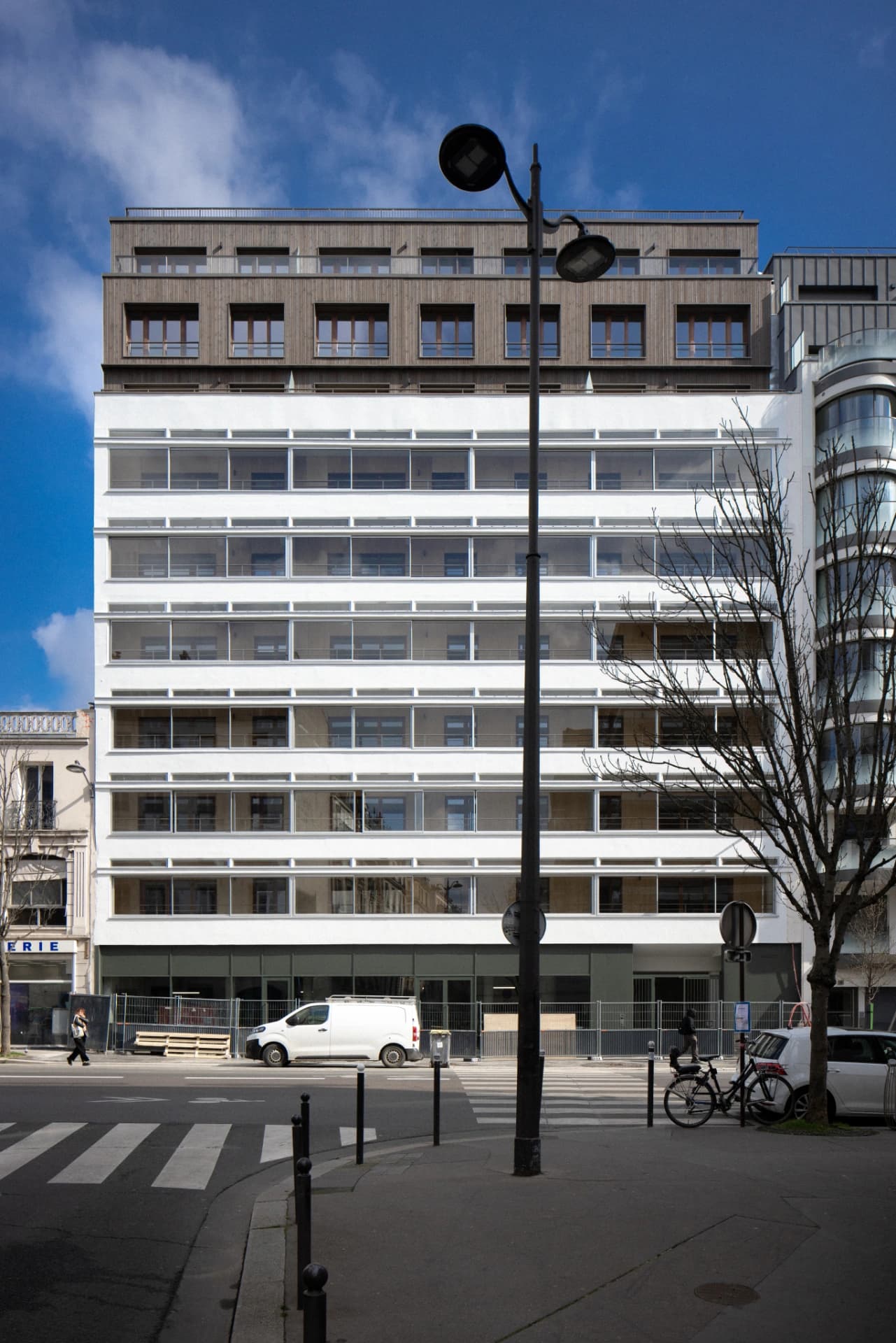 Un bâtiment moderne de plusieurs étages présente une façade blanche en partie basse et un revêtement en bois foncé en partie haute, sous un ciel bleu clair. Au premier plan, une rue urbaine animée avec un passage piéton, un lampadaire central, une camionnette blanche et un arbre dénudé.
