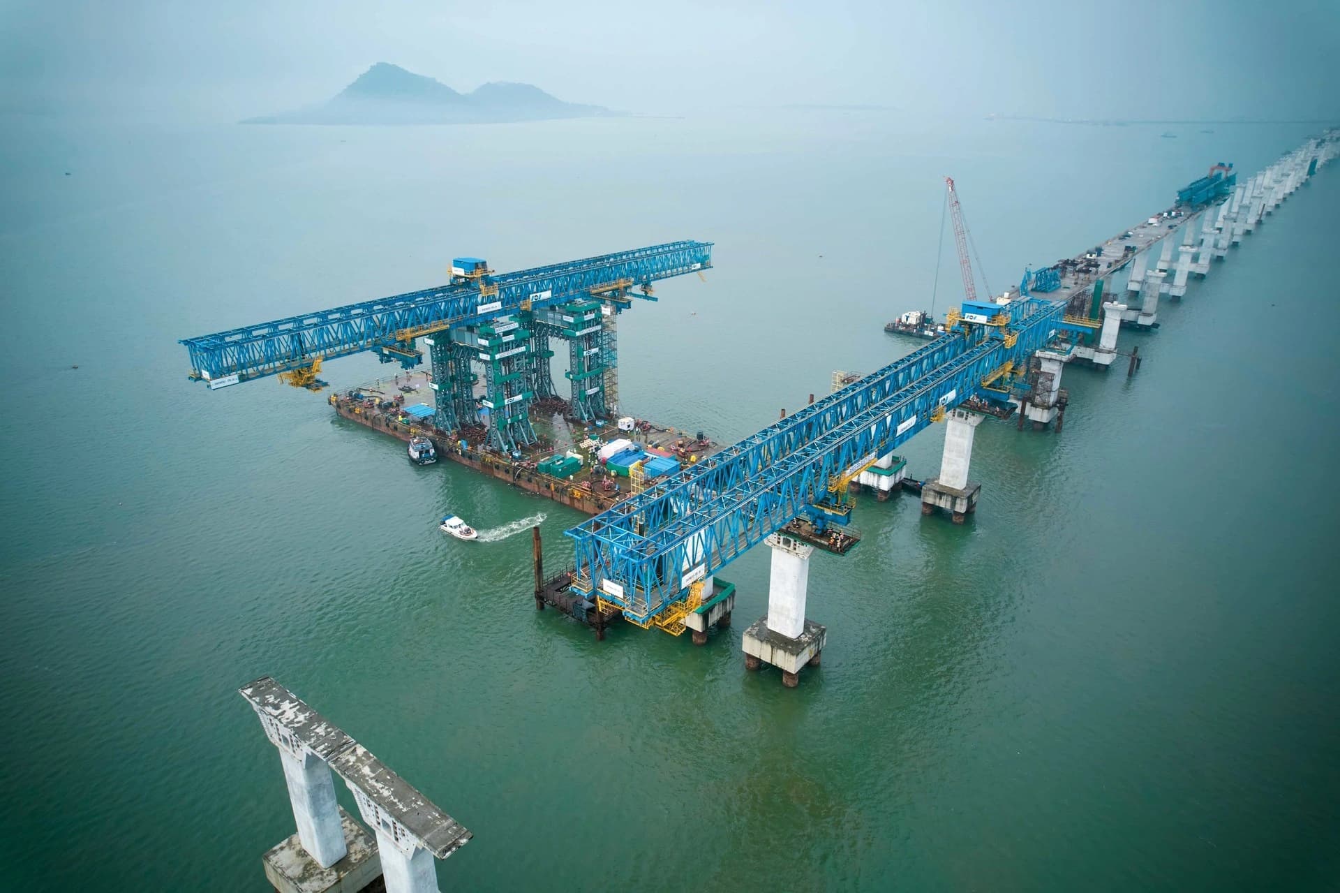 Vue aérienne du chantier d'un pont sur l'eau, montrant des poutres en acier bleu et des piliers en béton.