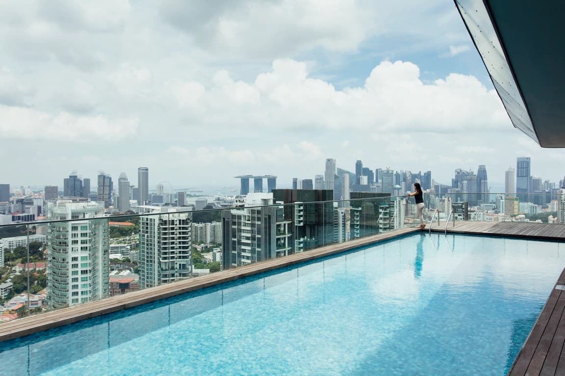 Une femme se tient au bord d'une piscine à débordement sur un toit, profitant de la vue imprenable sur les gratte-ciel de Singapour, dont le Marina Bay Sands, sous un ciel nuageux.