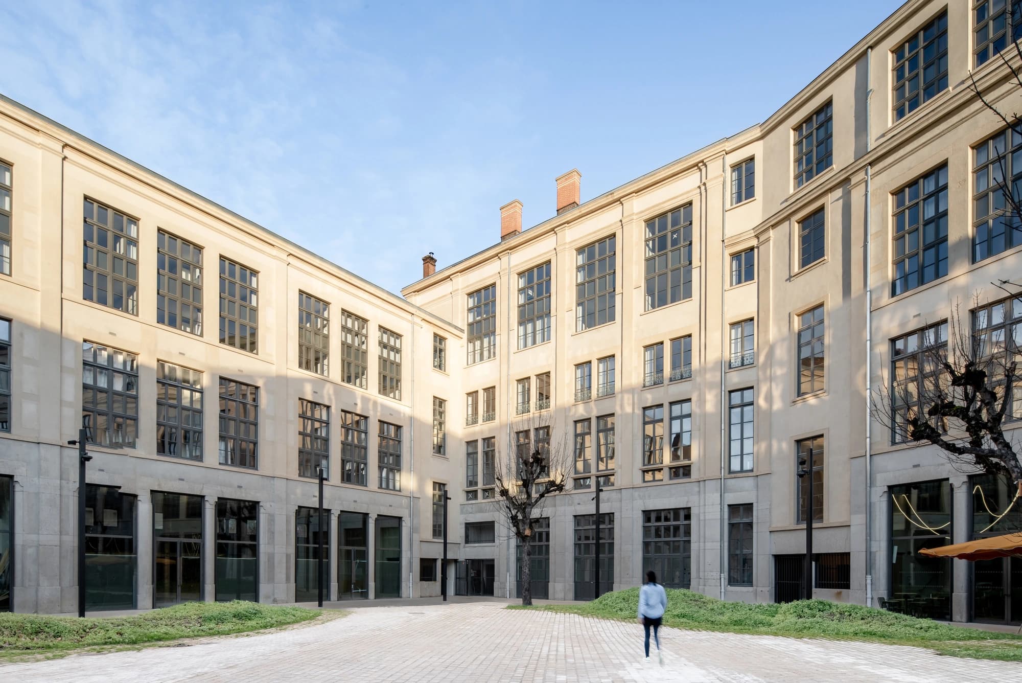 Un bâtiment en forme de U, de couleur beige clair et doté de nombreuses fenêtres à cadres sombres, entoure une cour pavée sous un ciel bleu dégagé. Une personne vêtue d'un haut bleu clair s'éloigne de l'appareil photo, avec des parterres de gazon vert et un arbre dénudé visibles au premier plan.