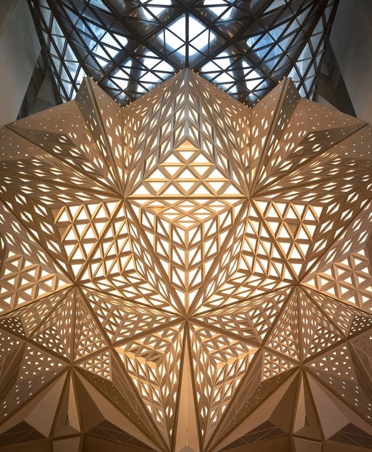 Intricate golden architectural ceiling featuring complex geometric panels illuminated by glowing triangular and diamond perforations, seen beneath a darker grid-patterned glass dome.